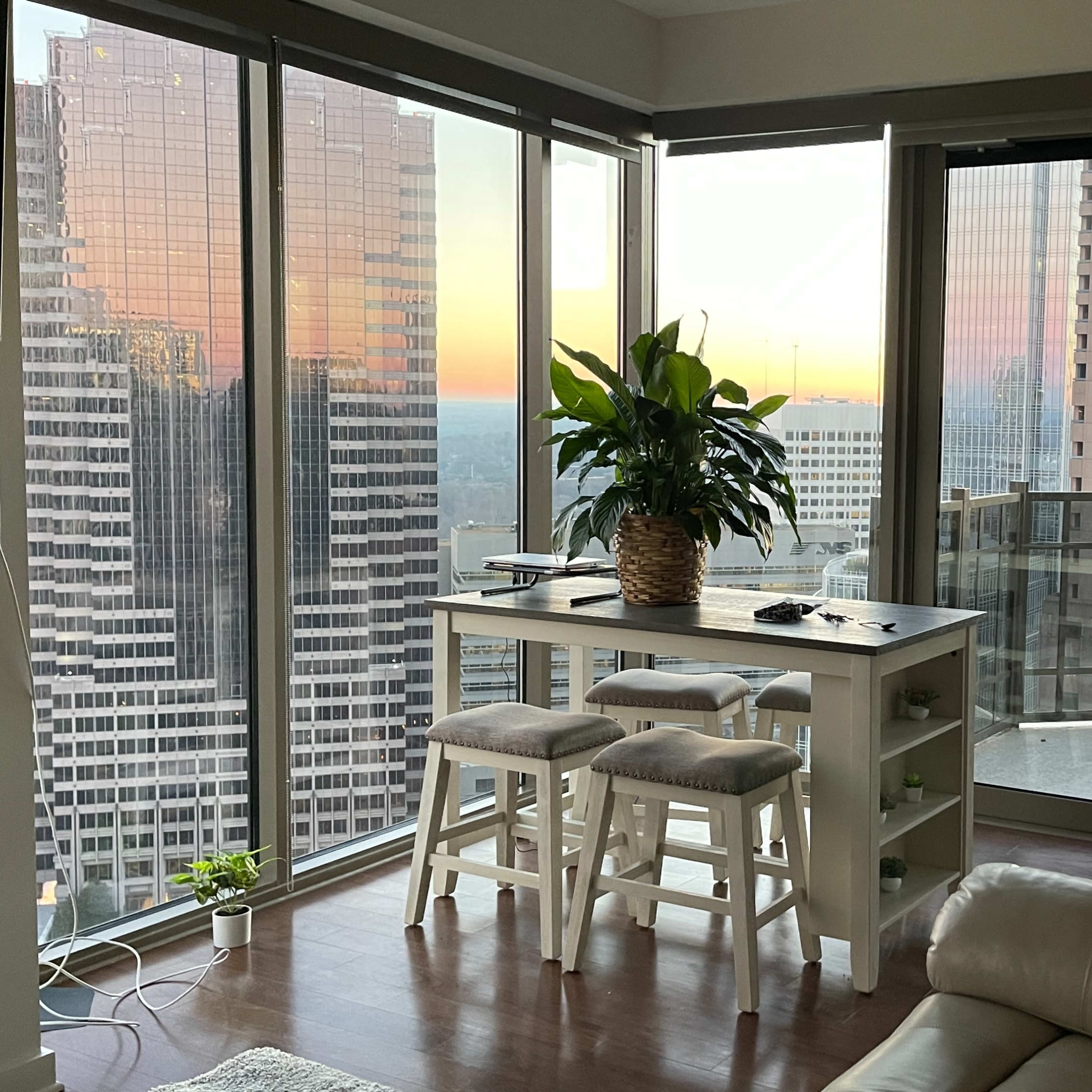 A modern apartment features a dining area with a plant, overlooking a city skyline at sunset through large glass windows.
