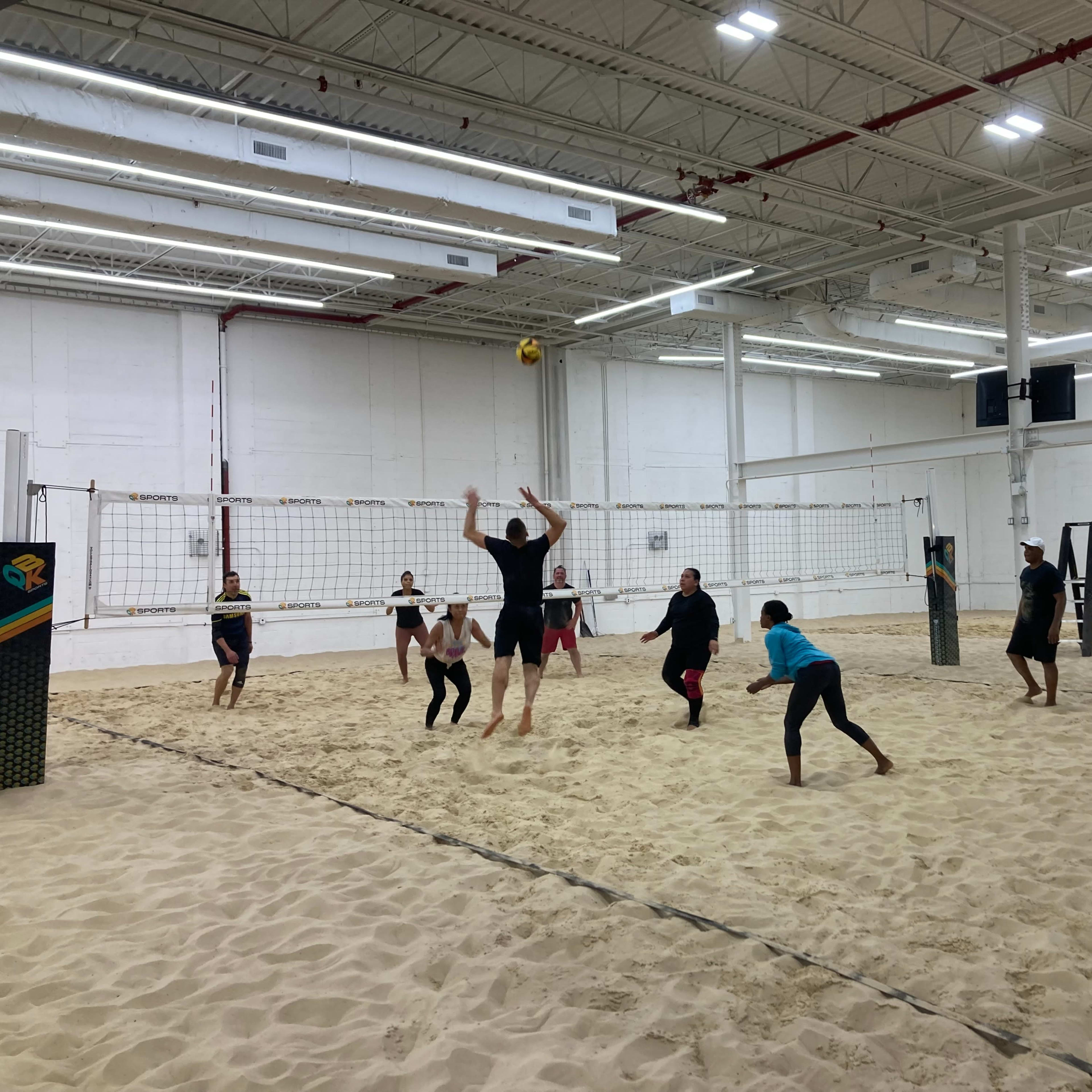 A group of six people play beach volleyball indoors on a sand court.