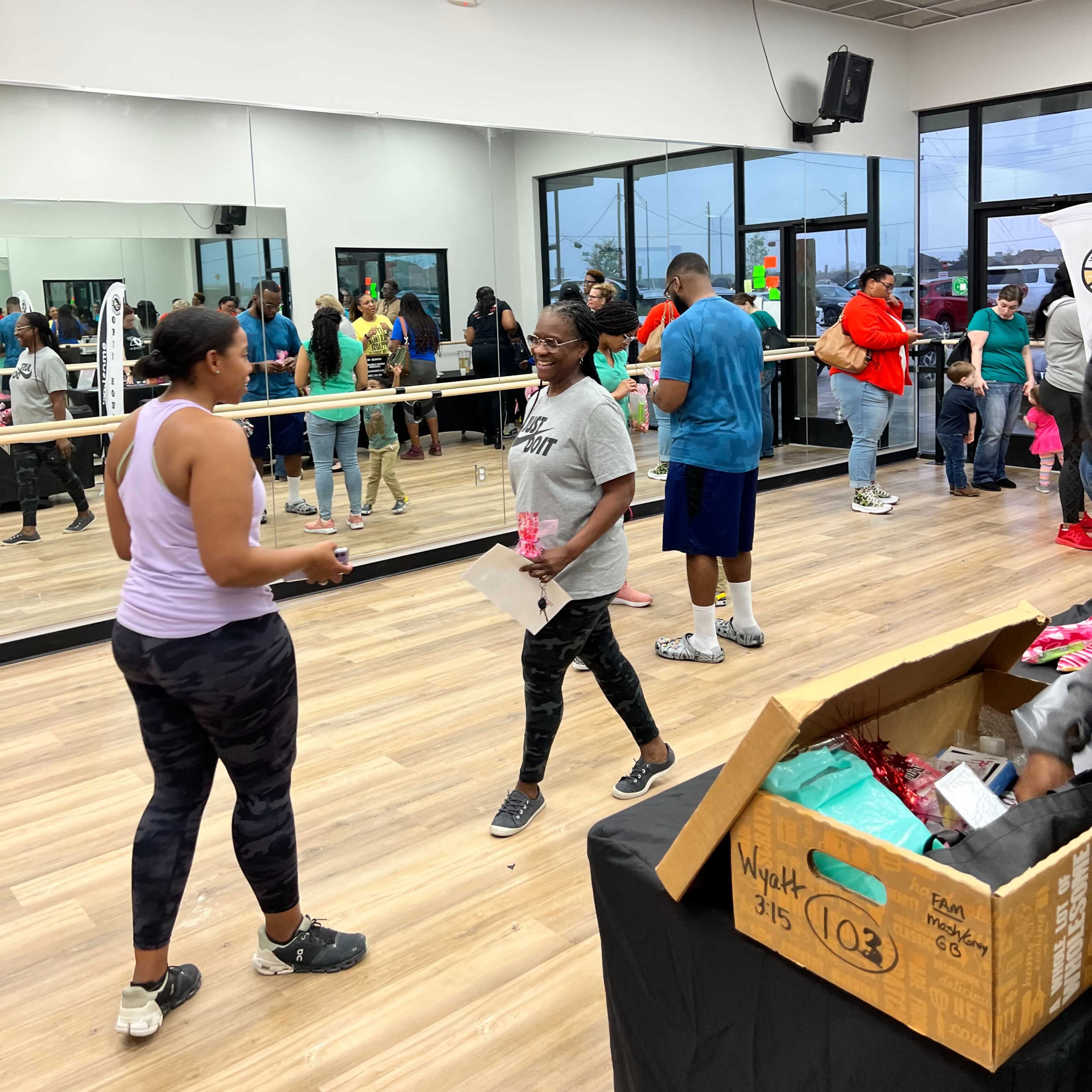 A group of people gather in a well-lit fitness studio with mirrors, some participating in an exercise class and others waiting around, while a box of supplies sits on a table.