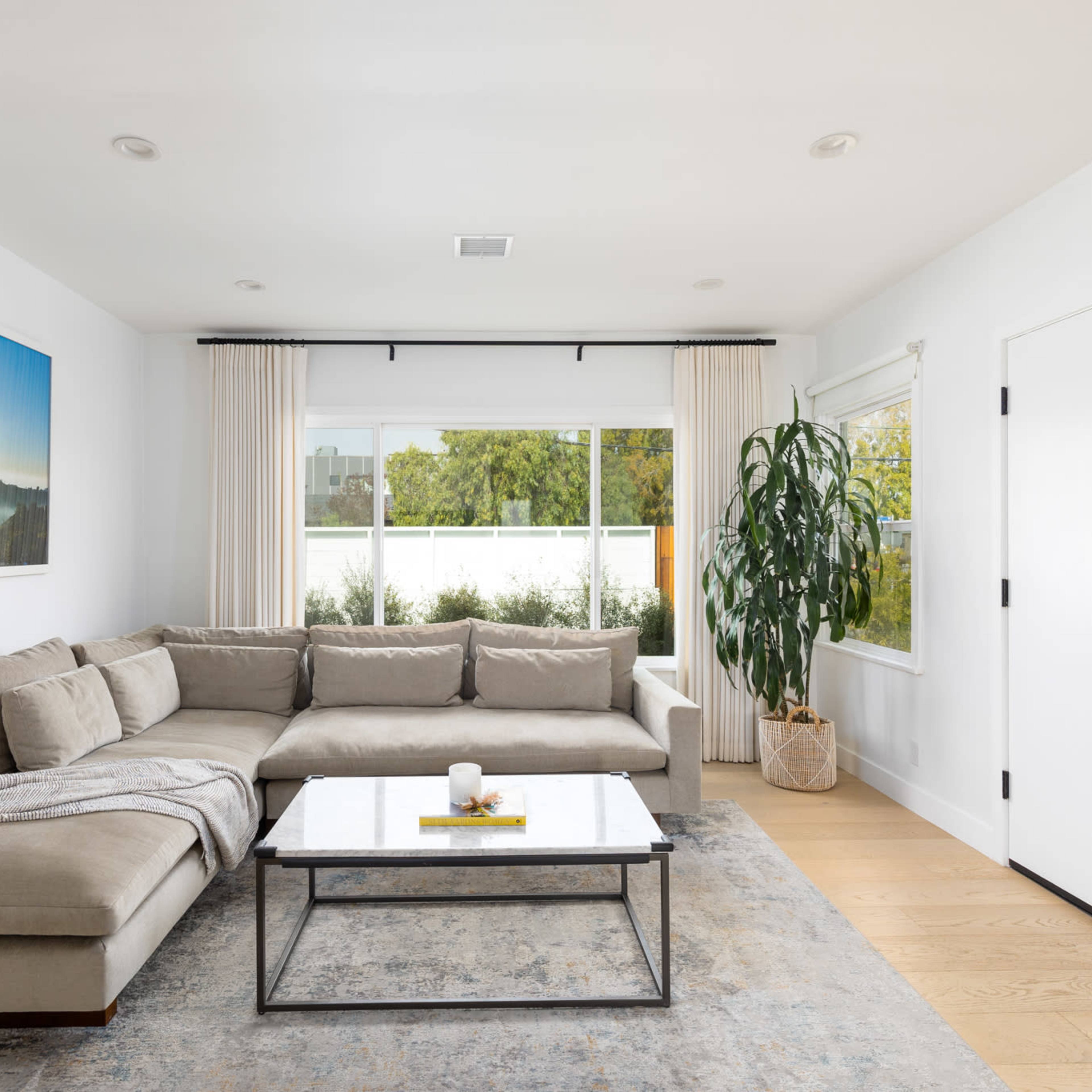 A modern living room features a large beige sectional sofa, a glass coffee table, and floor-to-ceiling windows with sheer curtains.