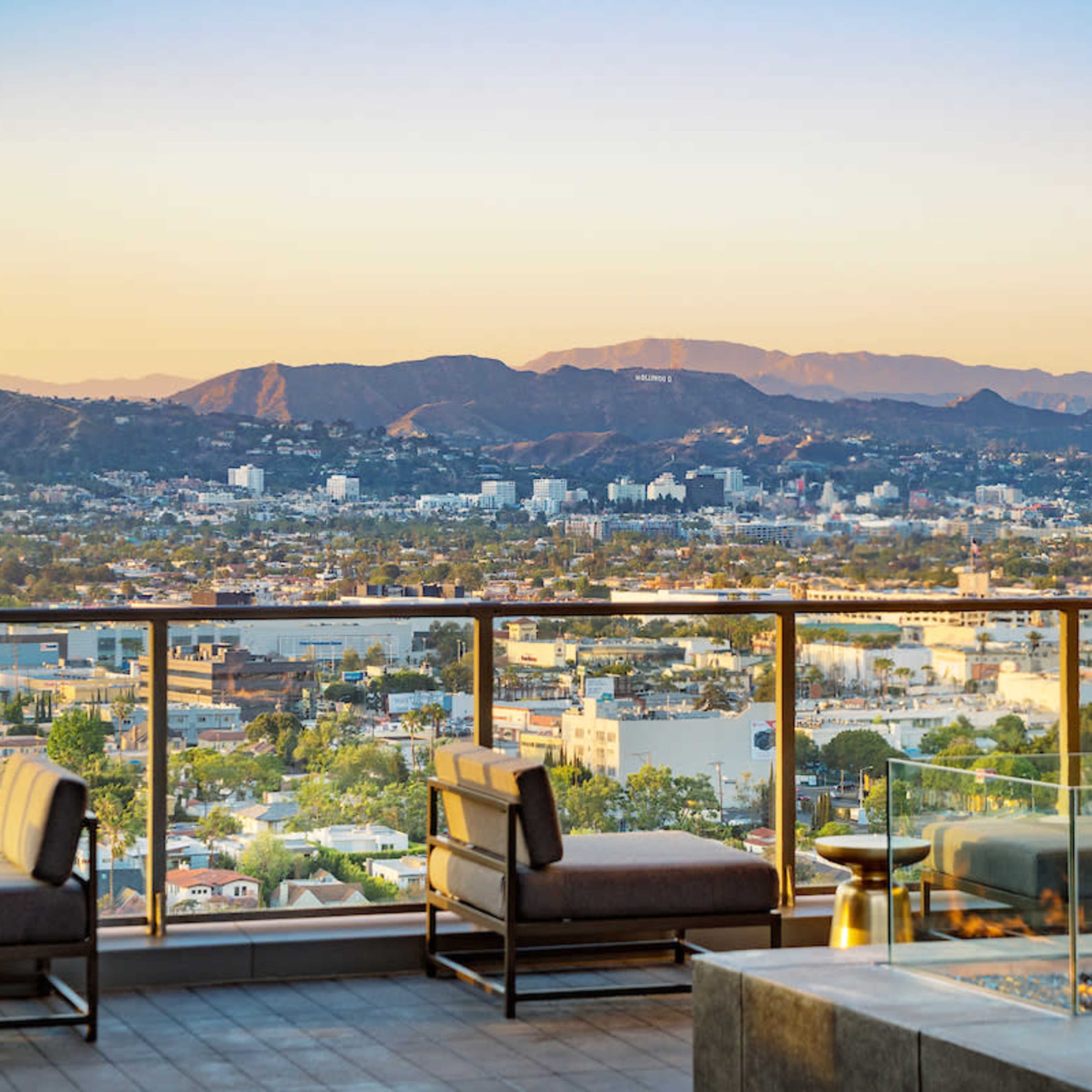 A rooftop terrace with modern seating and a fire pit overlooking a city skyline and mountains in the distance at sunset.