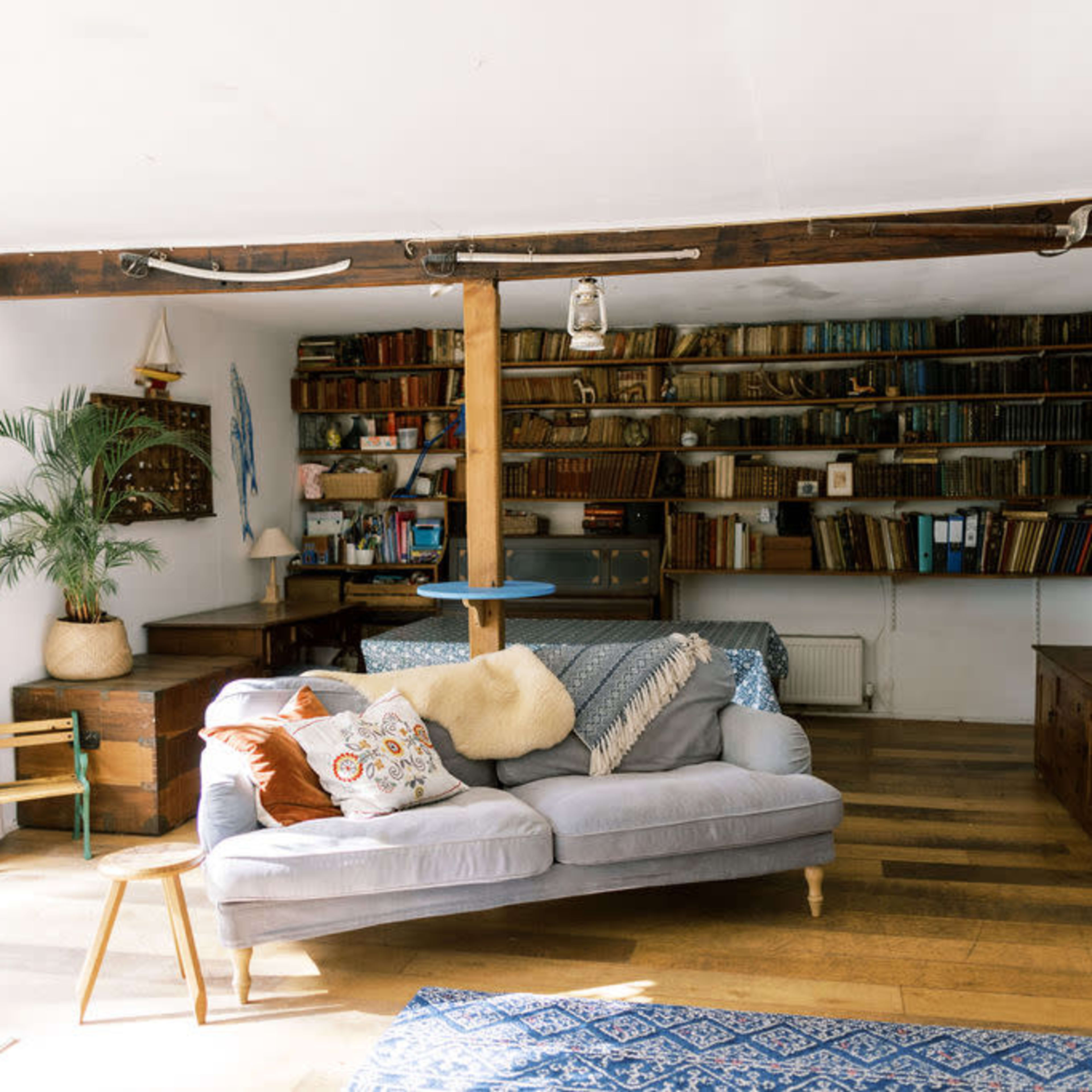 A cozy living room features a sofa with decorative cushions, a bookshelf filled with books, and a wooden beam supporting the ceiling.