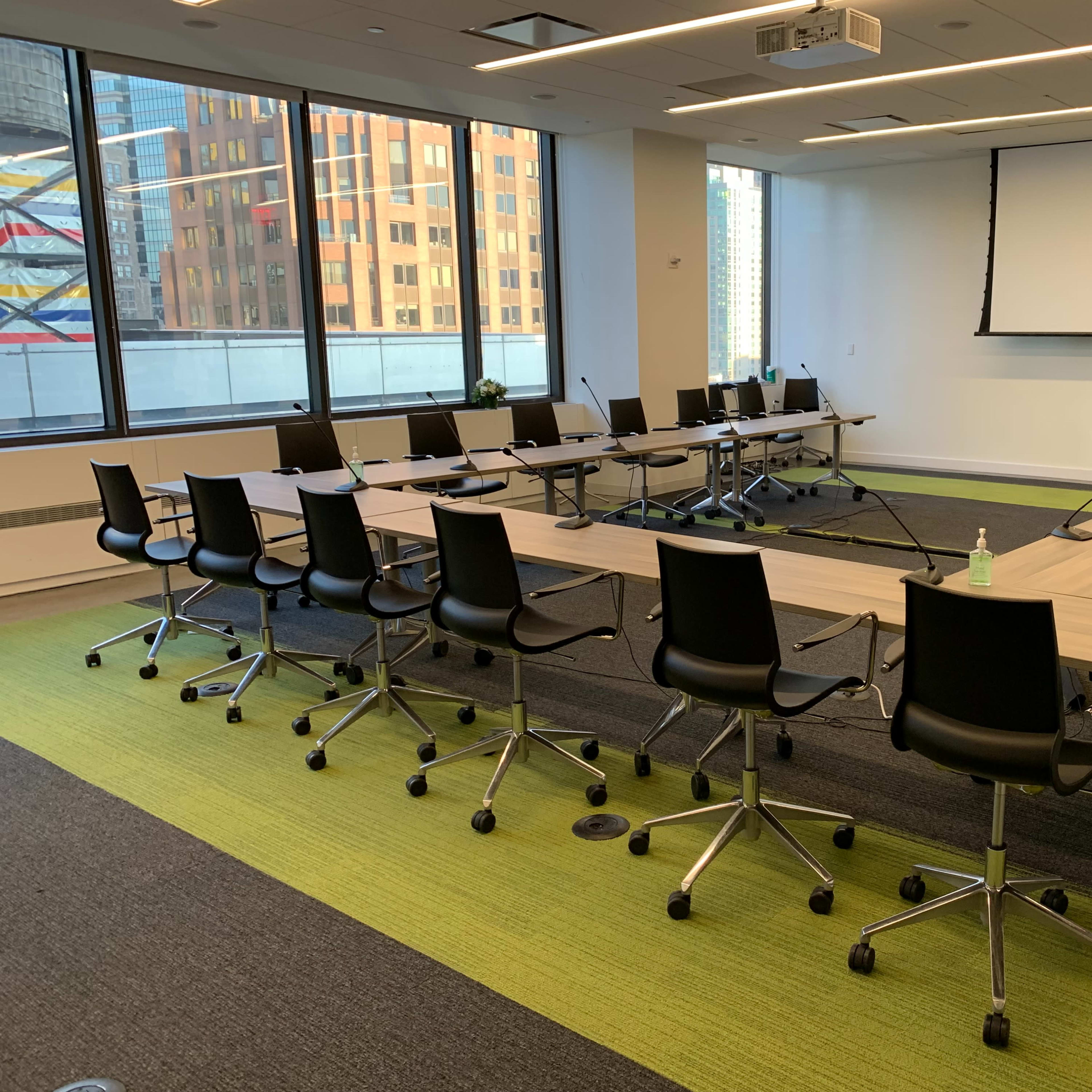 A large conference room with a long table surrounded by office chairs, large windows revealing a cityscape, and a projector screen mounted on the wall.