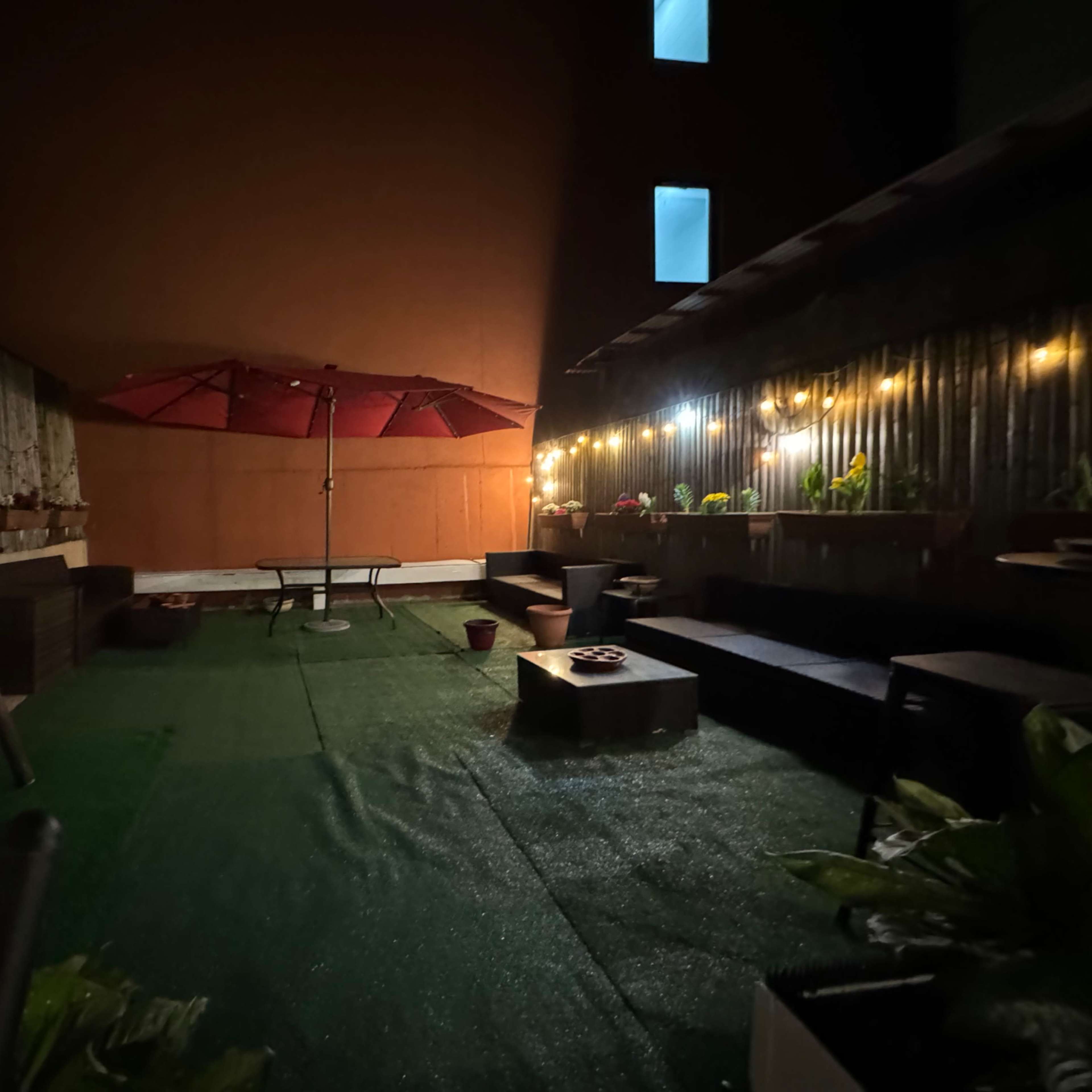A dimly lit outdoor terrace with green artificial grass, a central table, a red umbrella, and string lights illuminating the surrounding plants and seating areas.
