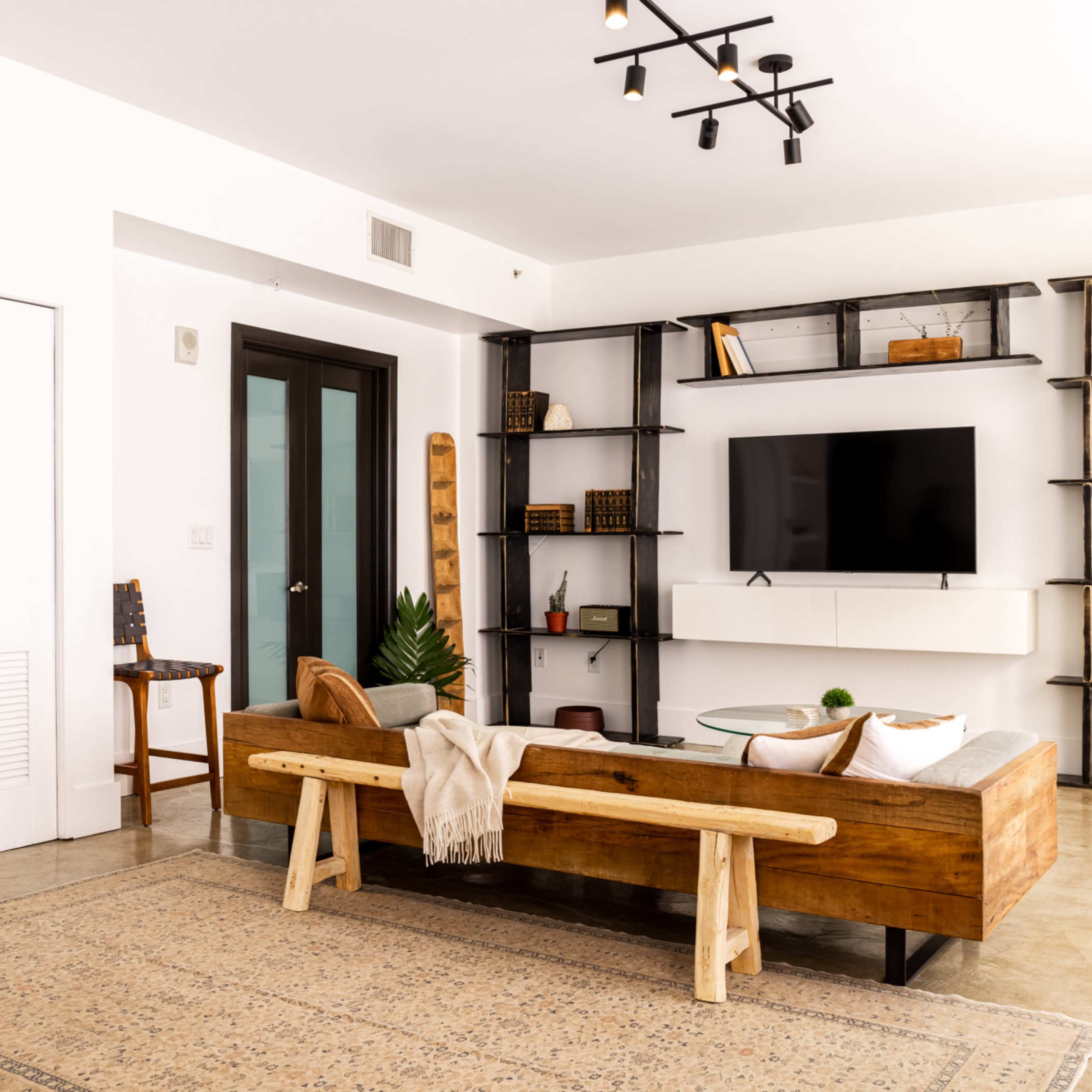 A modern living room features a wooden sofa, a coffee table, and wall shelves displaying decorative items and a television.