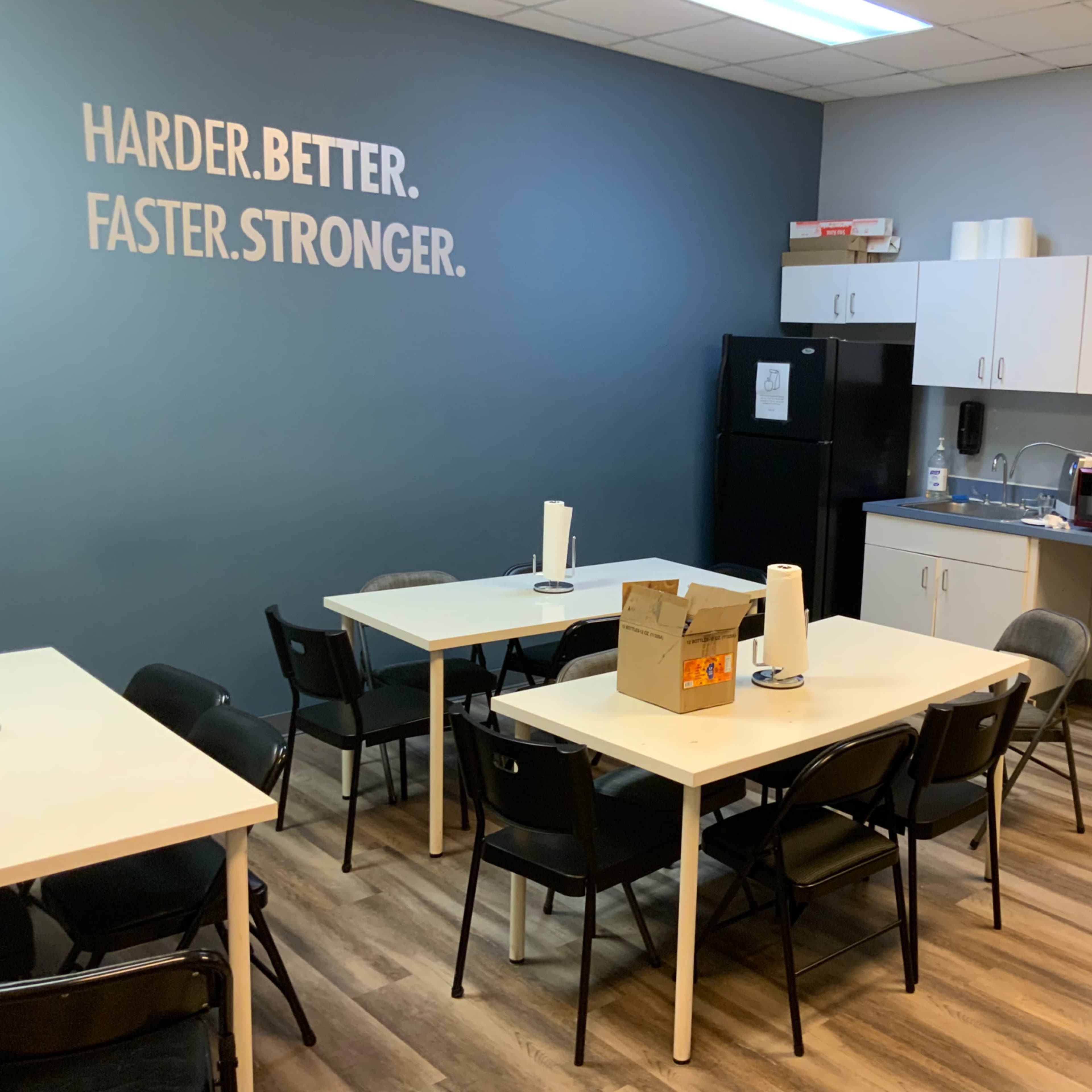 A break room features multiple tables and chairs, a refrigerator, a microwave, and a wall displaying the text "HARDER. BETTER. FASTER. STRONGER."