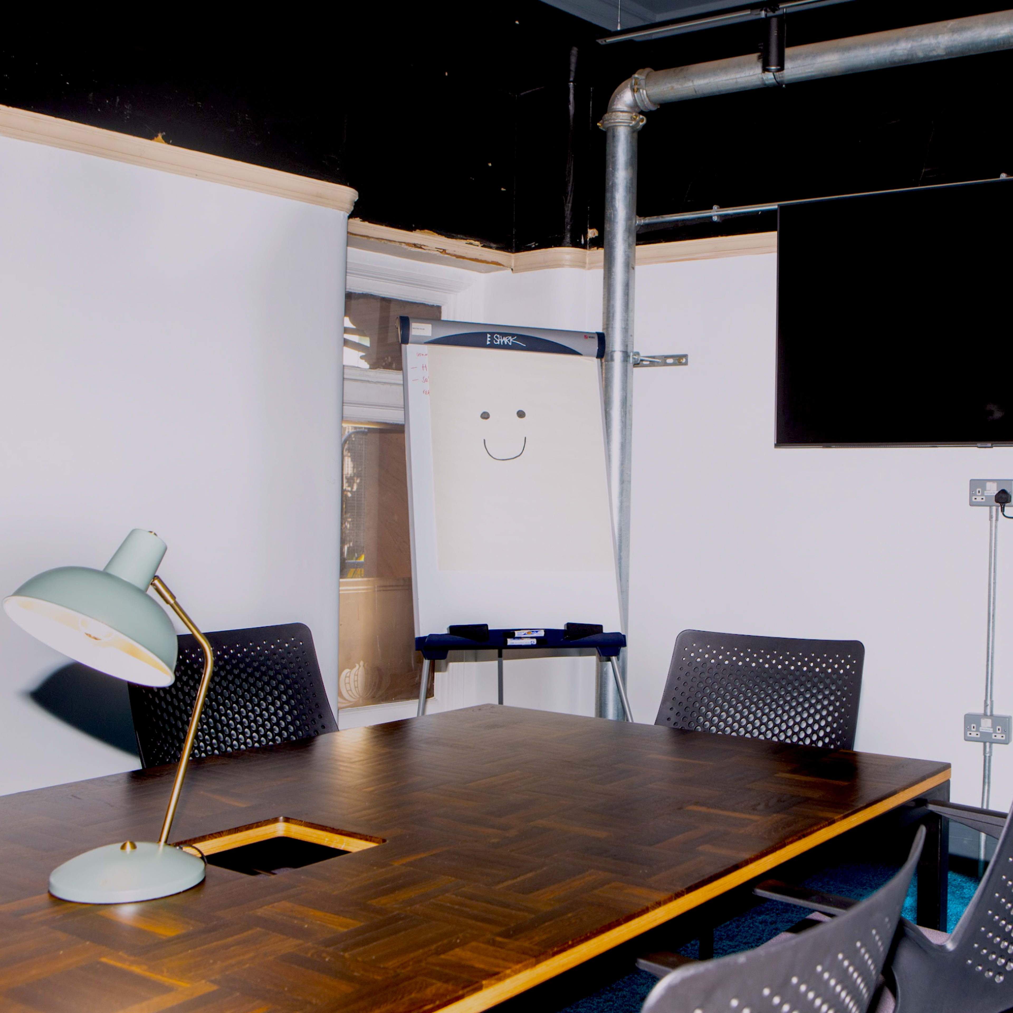 A conference room features a wooden table surrounded by black chairs, with a whiteboard displaying a smiley face and a television mounted on the wall.