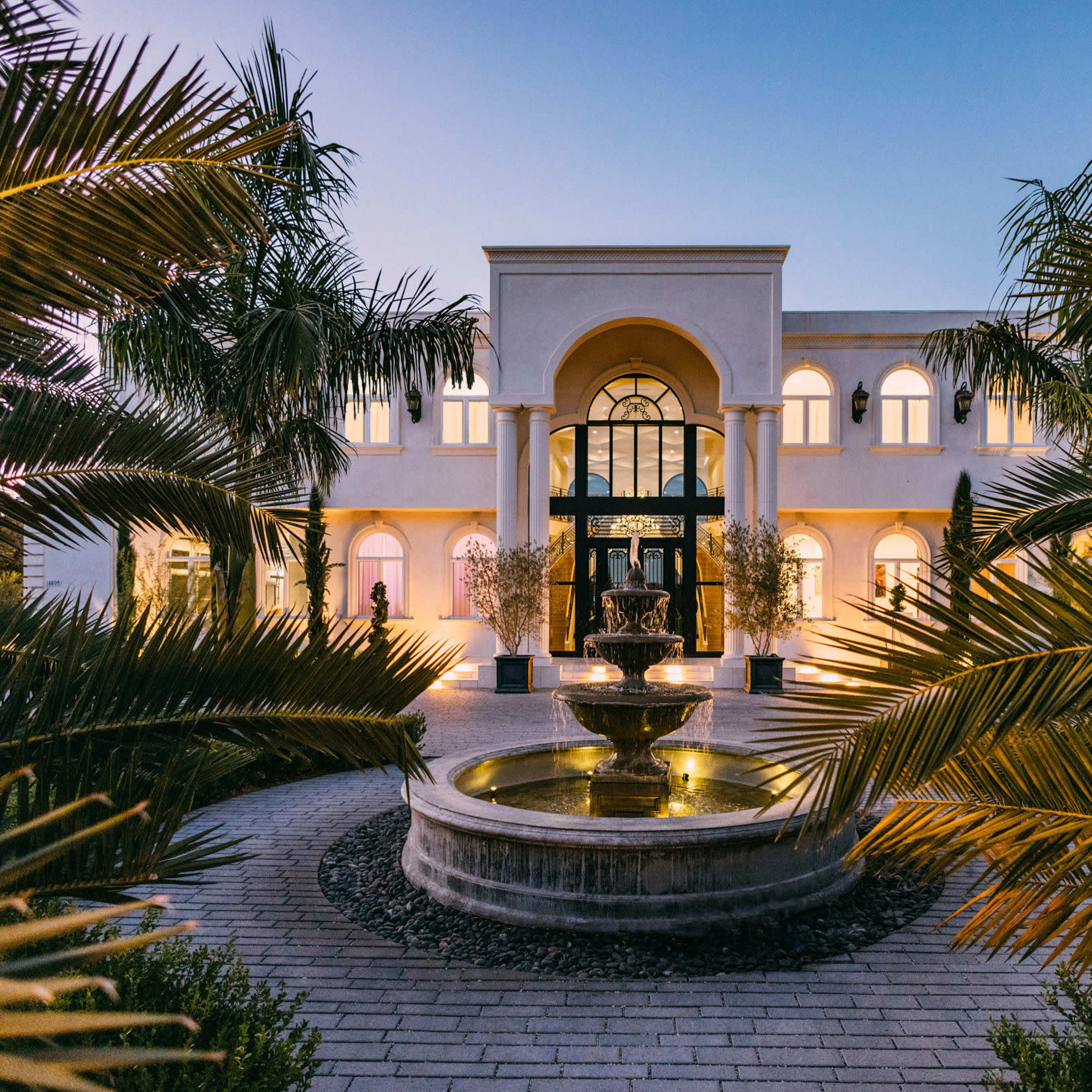A grand building with arched windows and a central fountain is surrounded by palm trees in a landscaped area.