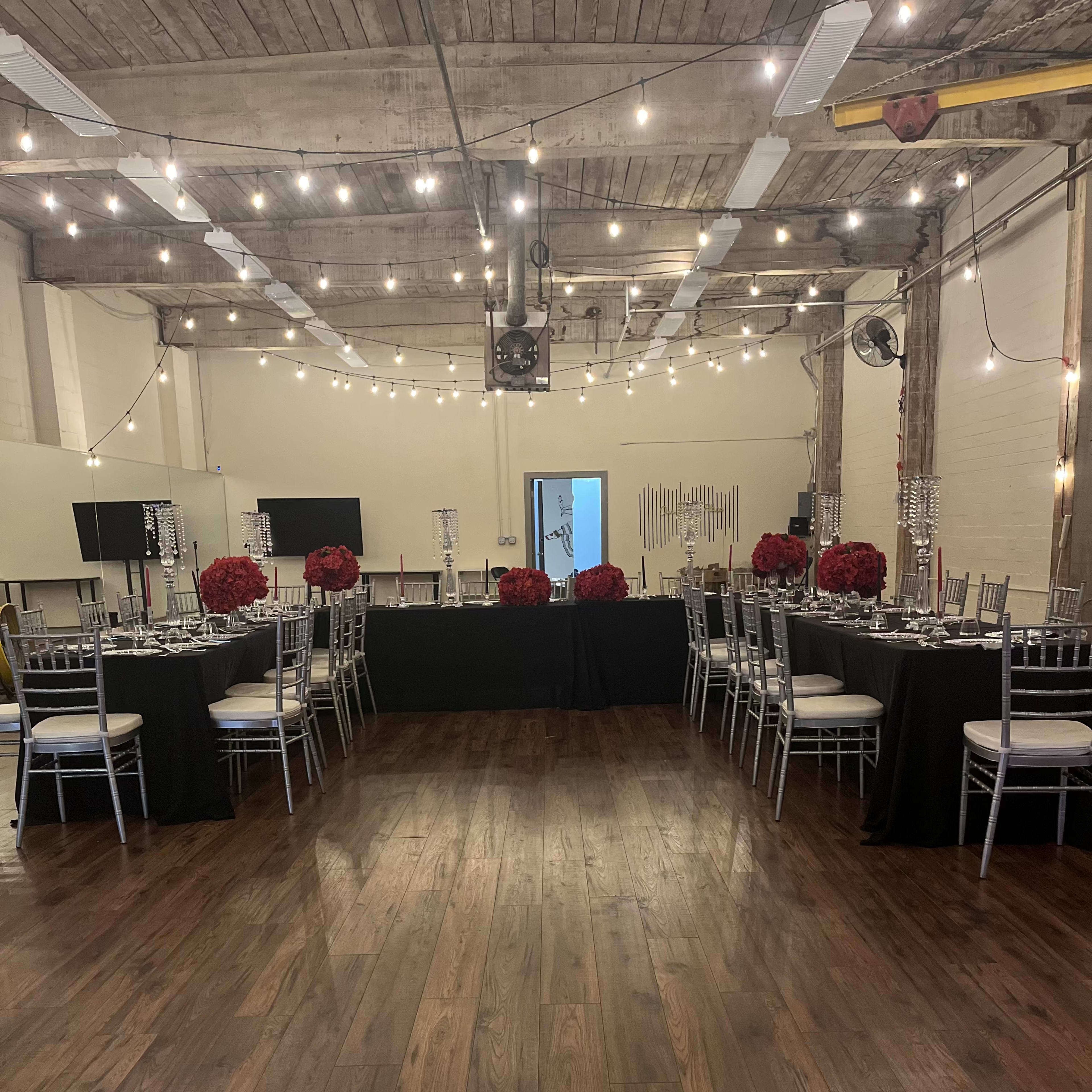 A spacious event venue features tables set with dark tablecloths and elegant centerpieces, surrounded by wooden flooring and string lights overhead.