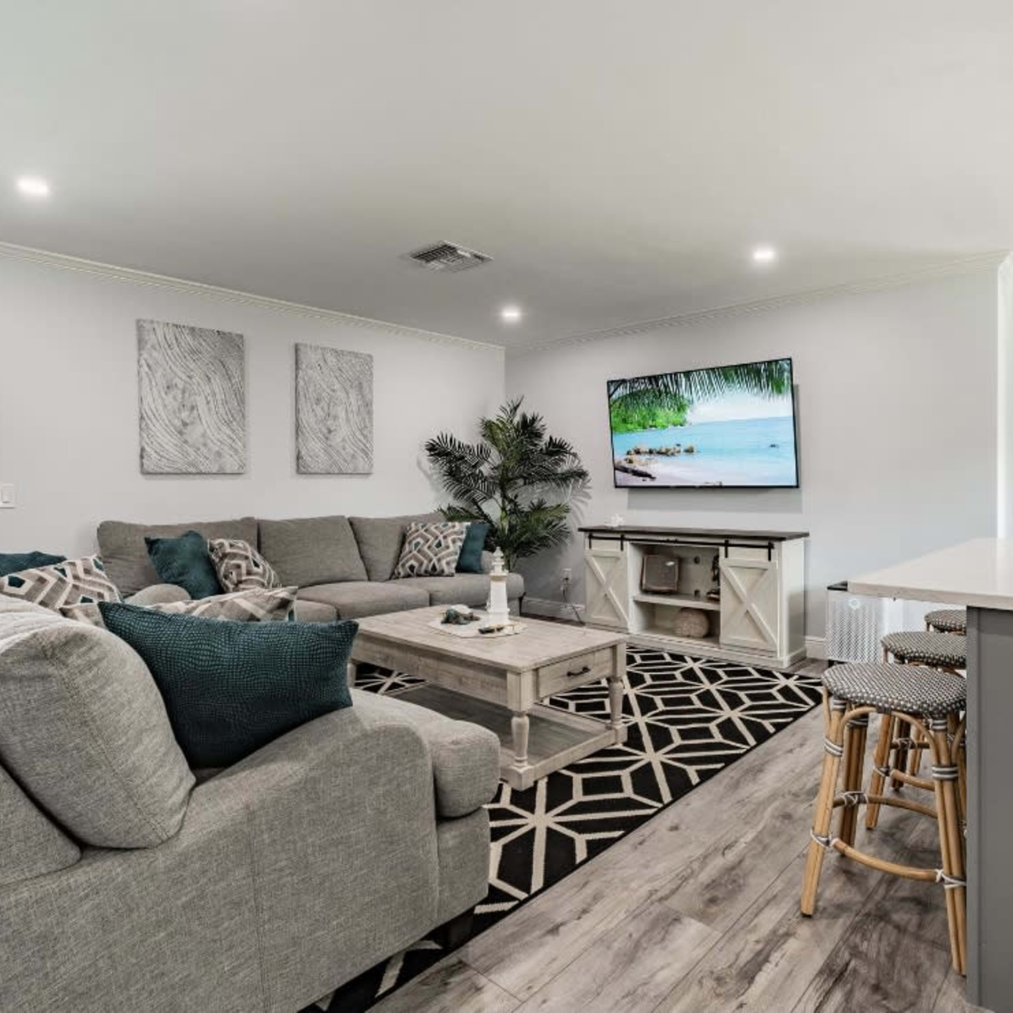A modern living room features a large gray sectional sofa, a coffee table, and a flat-screen TV displaying a scenic beach image, with a kitchen area visible in the background.