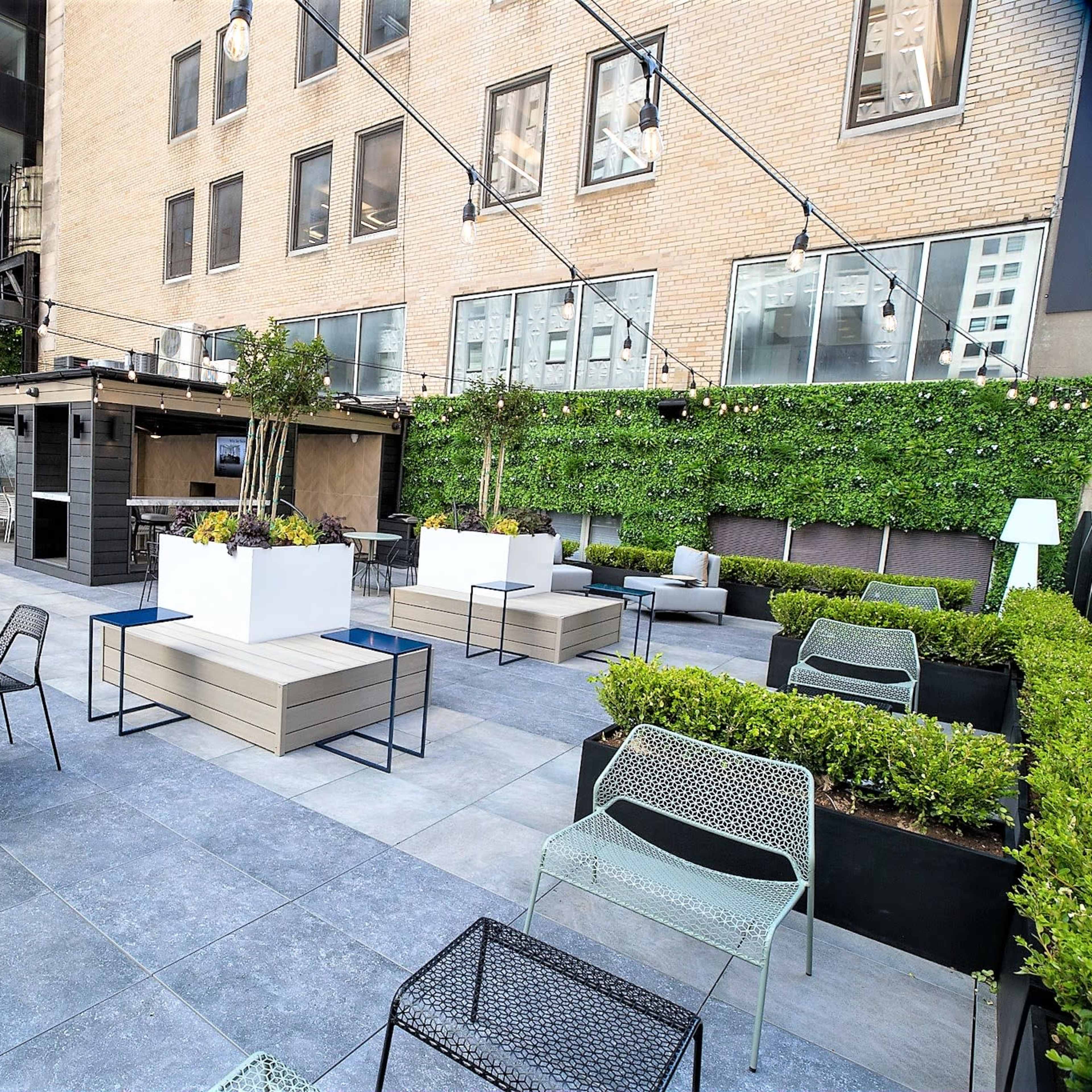 A modern rooftop patio area with tables and chairs, surrounded by greenery and partially enclosed by buildings.