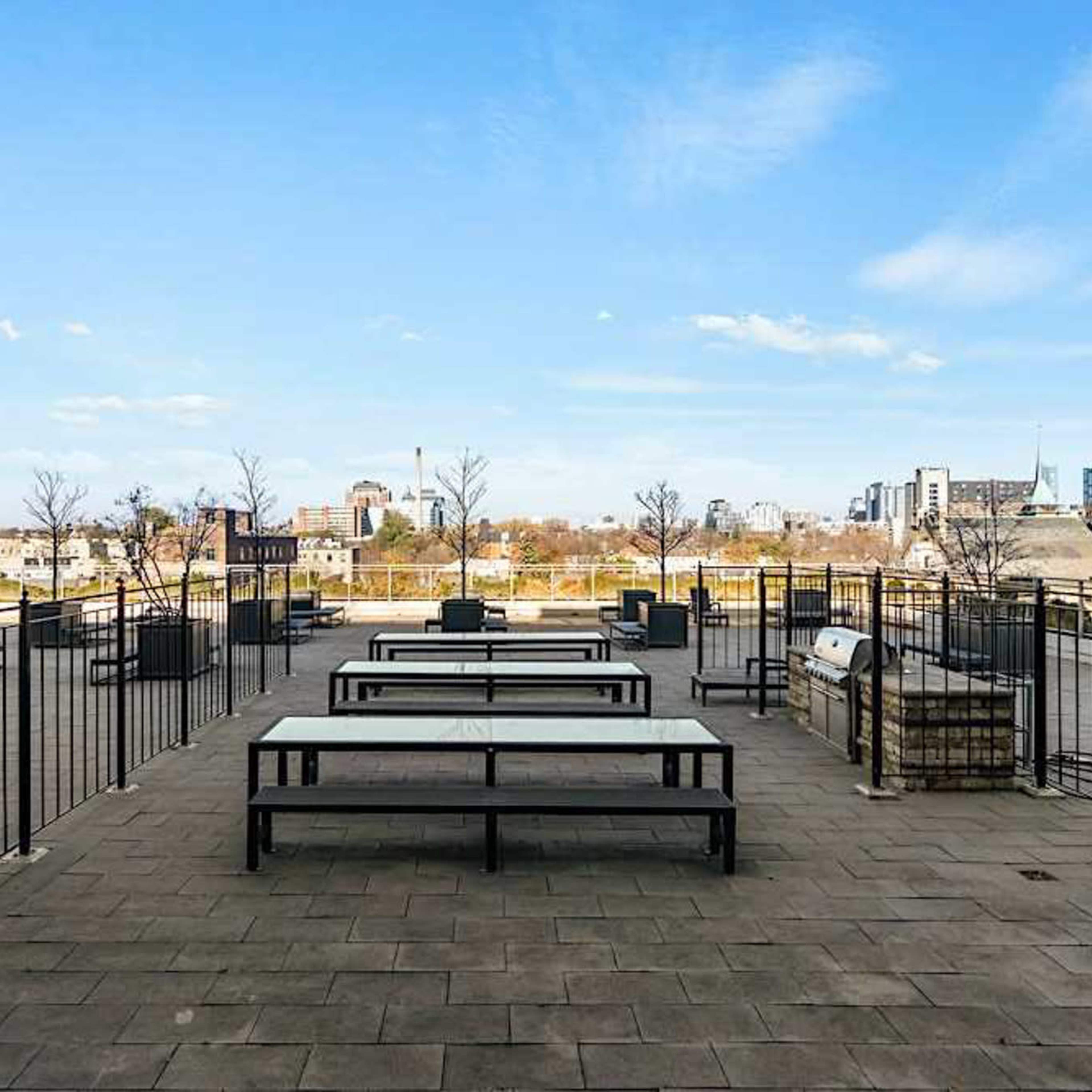 An empty rooftop terrace with metal railings, several picnic tables, and a view of a city skyline in the background.