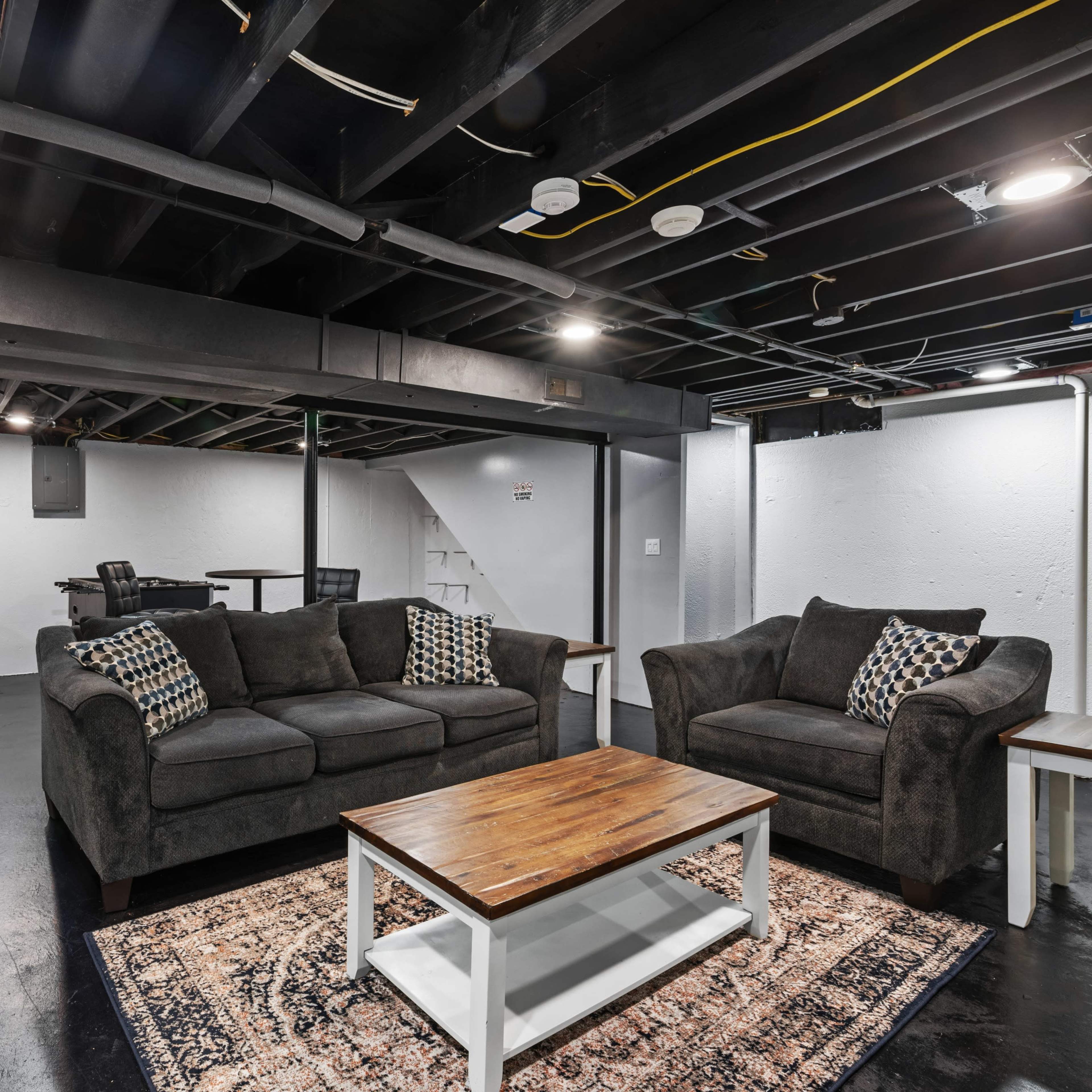 A finished basement with black ceilings, a coffee table, two dark sofas with pillows, and a small dining area.
