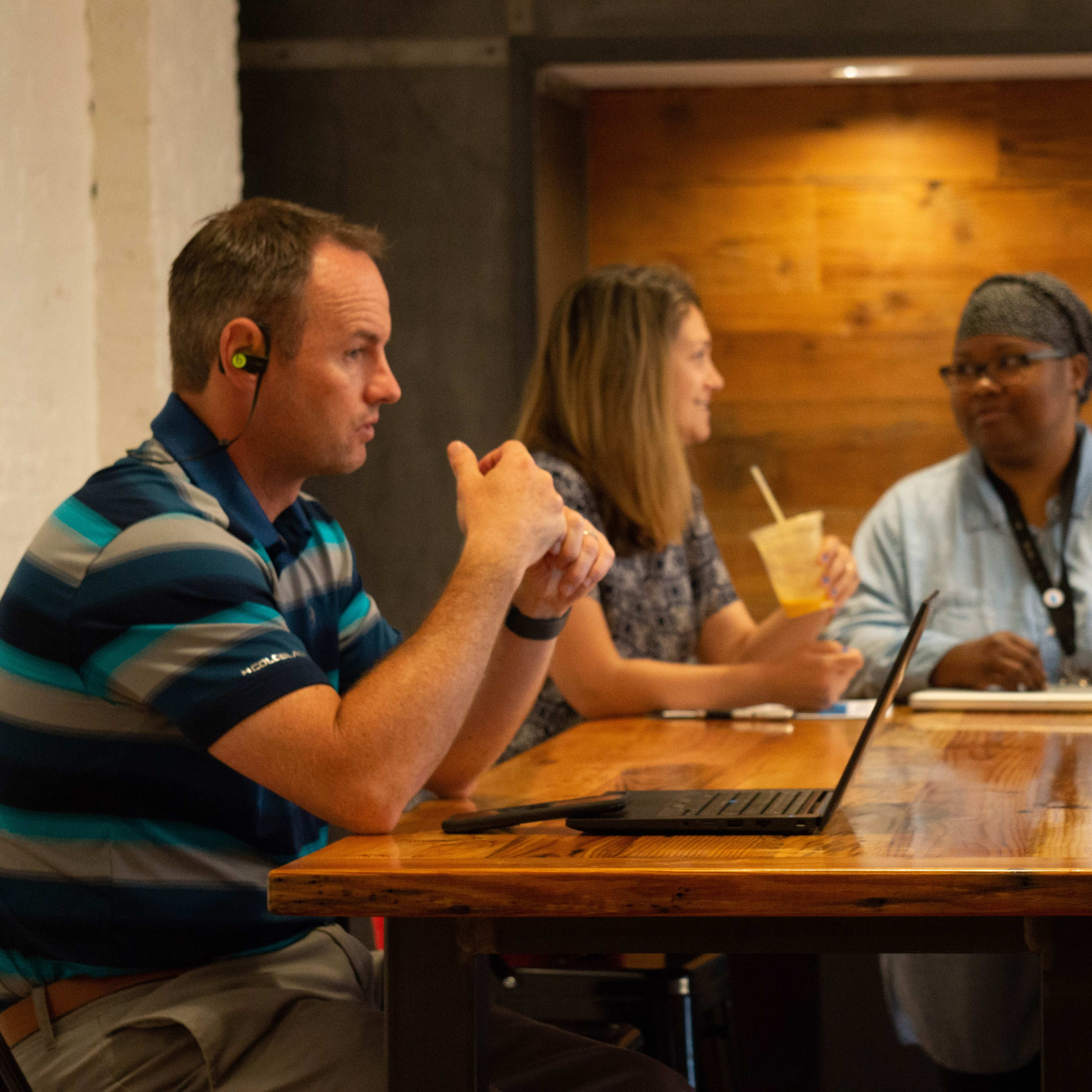 Three people are sitting at a wooden table in a cafe, with one person using a laptop and the others engaged in conversation.