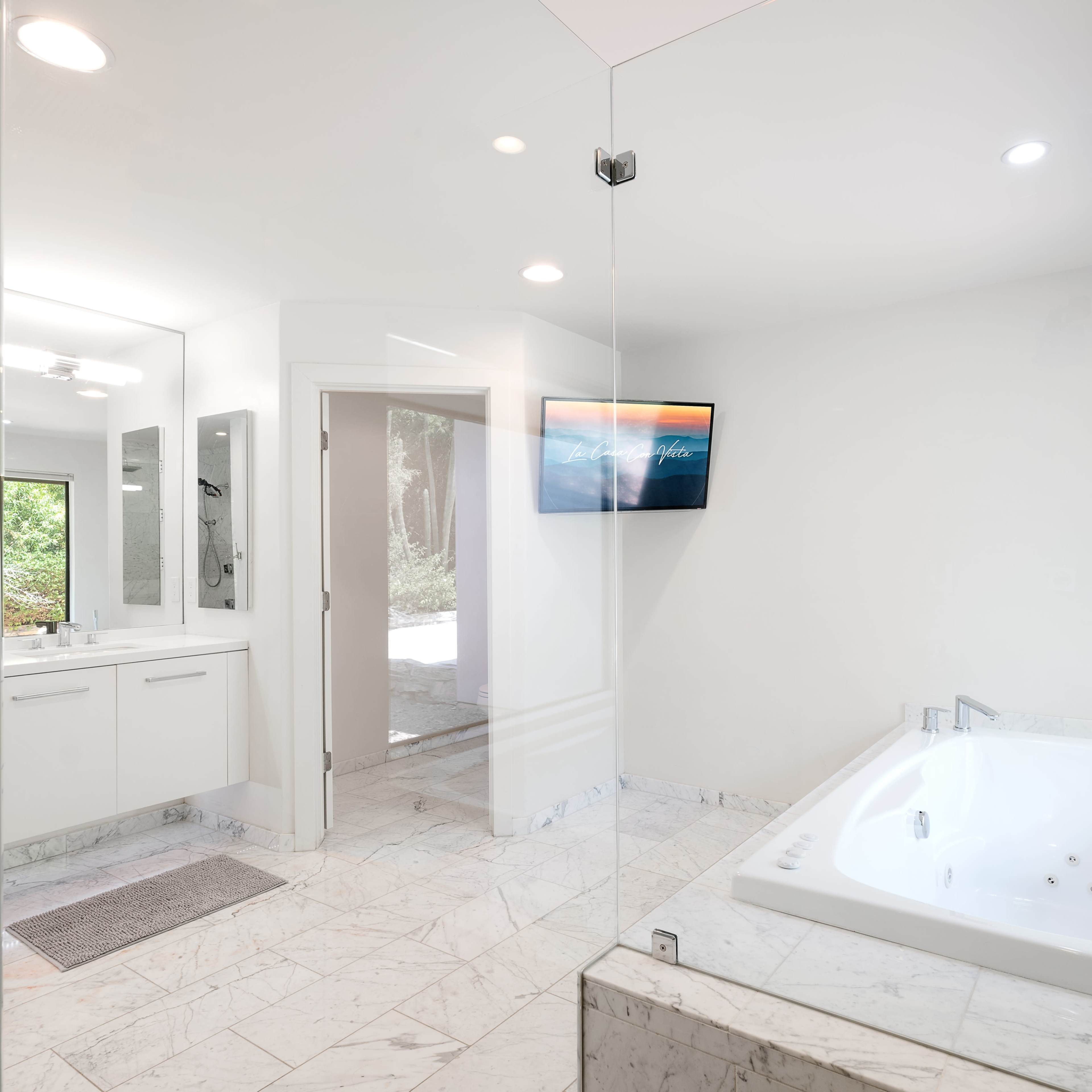 A modern bathroom with a glass shower, a white bathtub, a vanity with mirror, and a wall-mounted television.