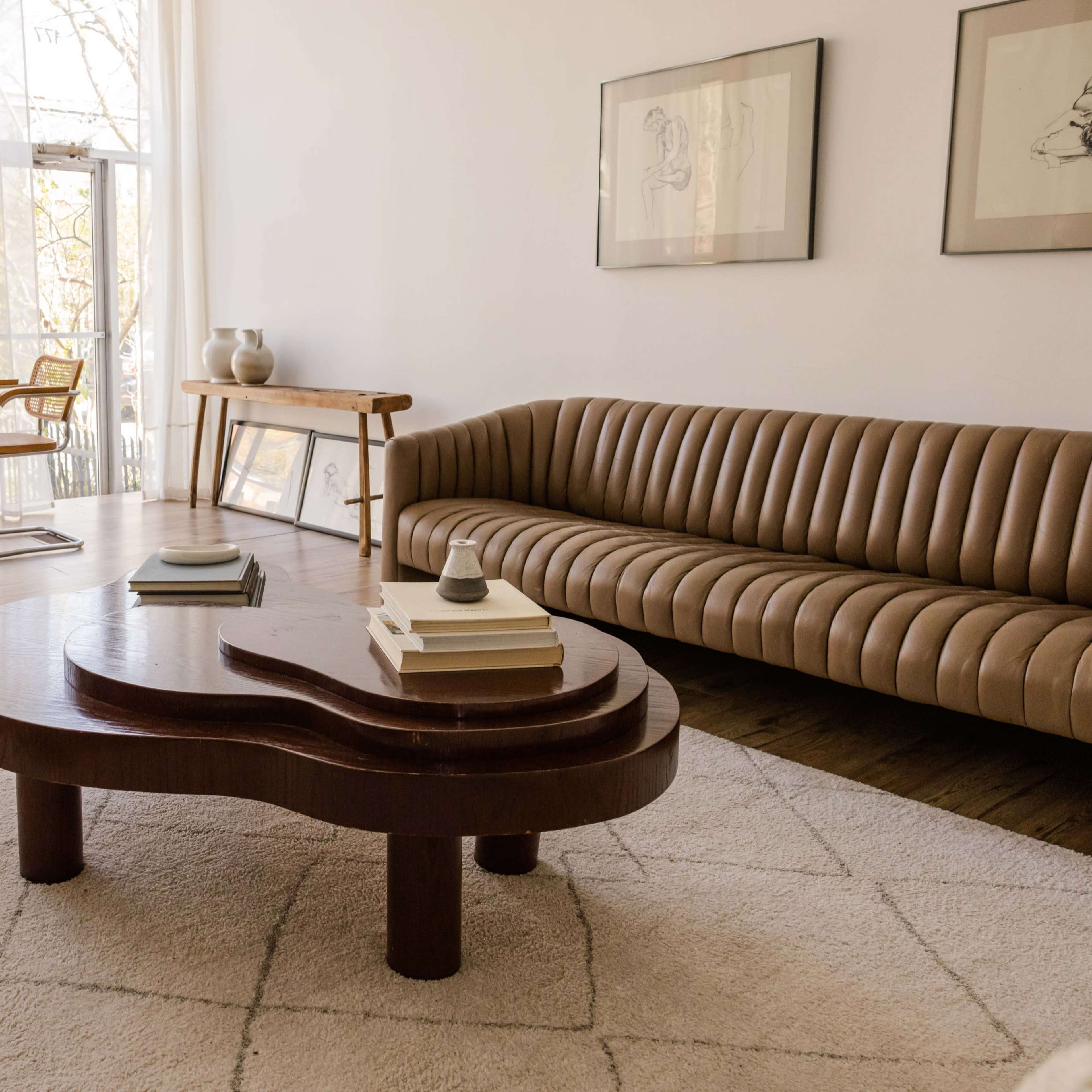 A modern living room featuring a curved brown sofa, a wooden coffee table with books, a minimalist chair, and a dining area with a round table and chairs, illuminated by natural light from large windows.