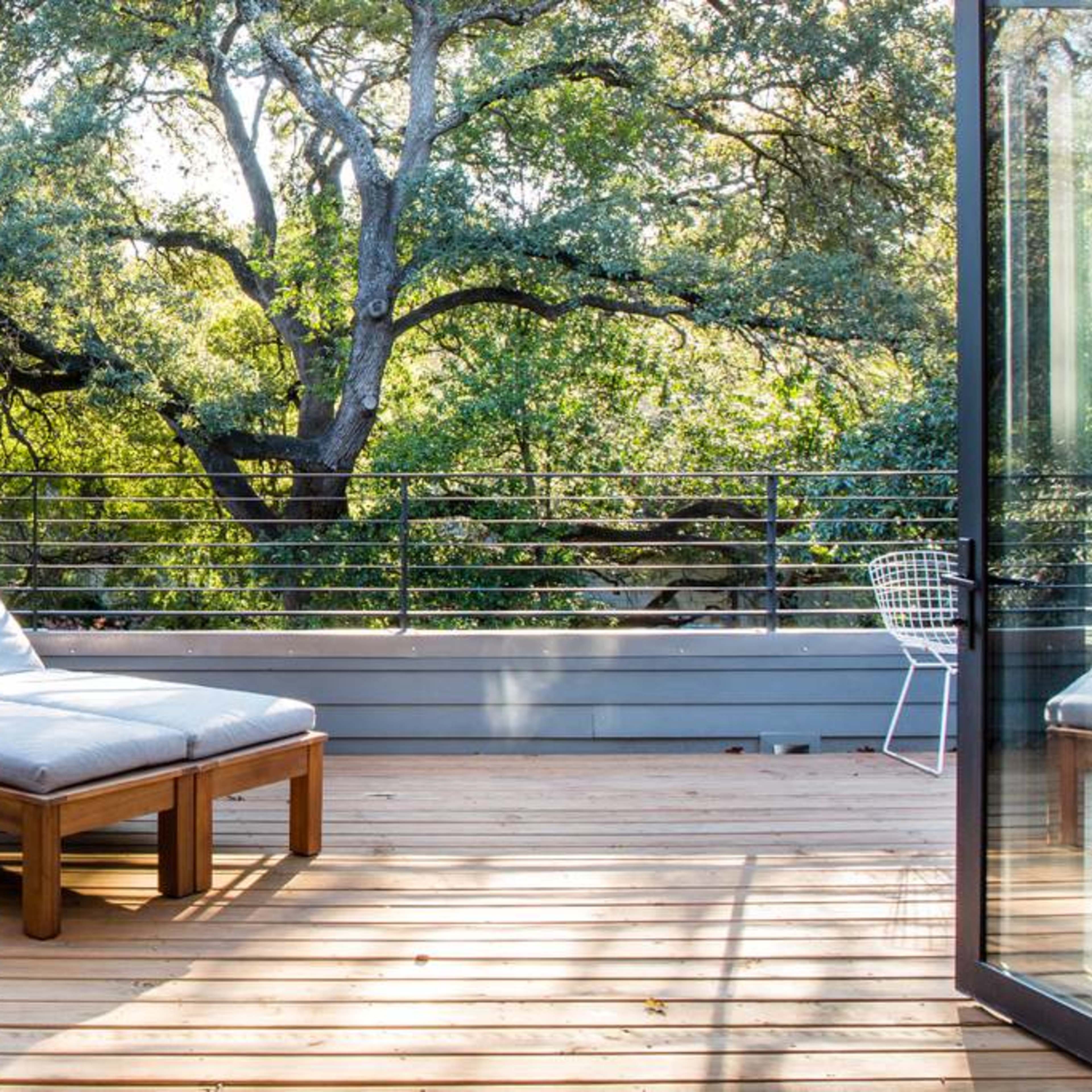 A wooden balcony features a lounge chair and a small chair, overlooking lush trees and greenery.