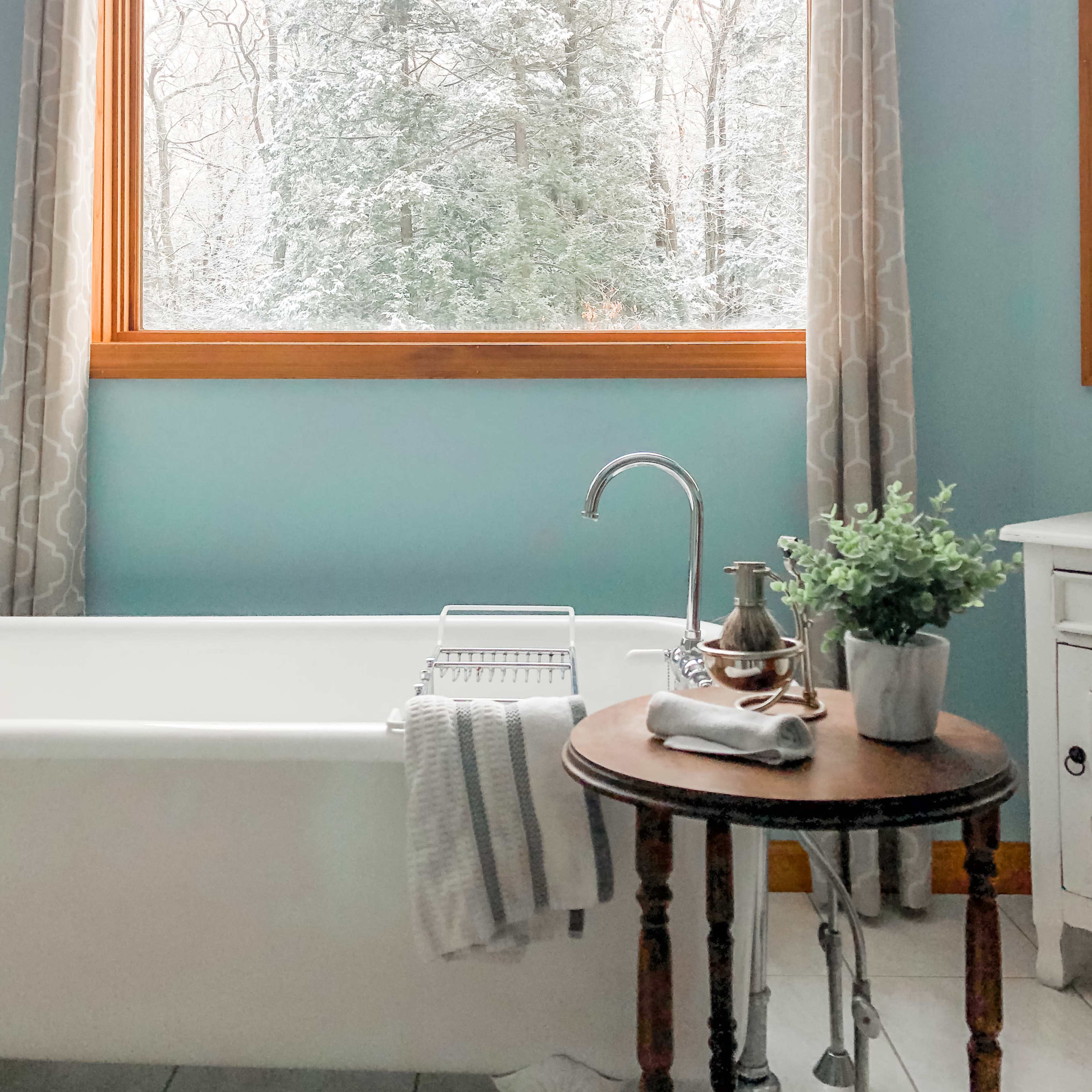 A bathroom features a freestanding bathtub beside a wooden table, with a snowy landscape visible through a large window.