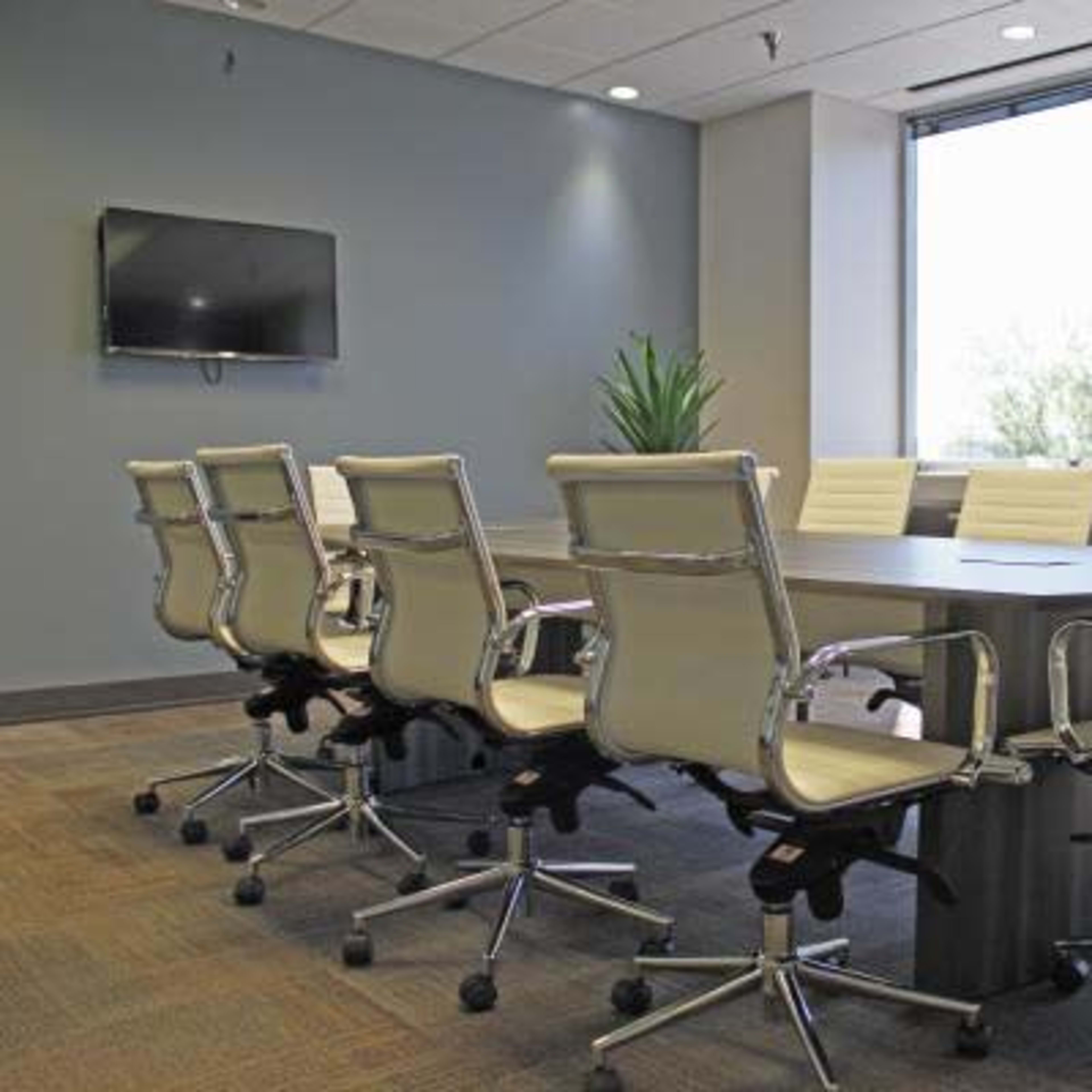 A modern conference room features a large table surrounded by ergonomic chairs, with a television mounted on the wall and windows offering daylight.
