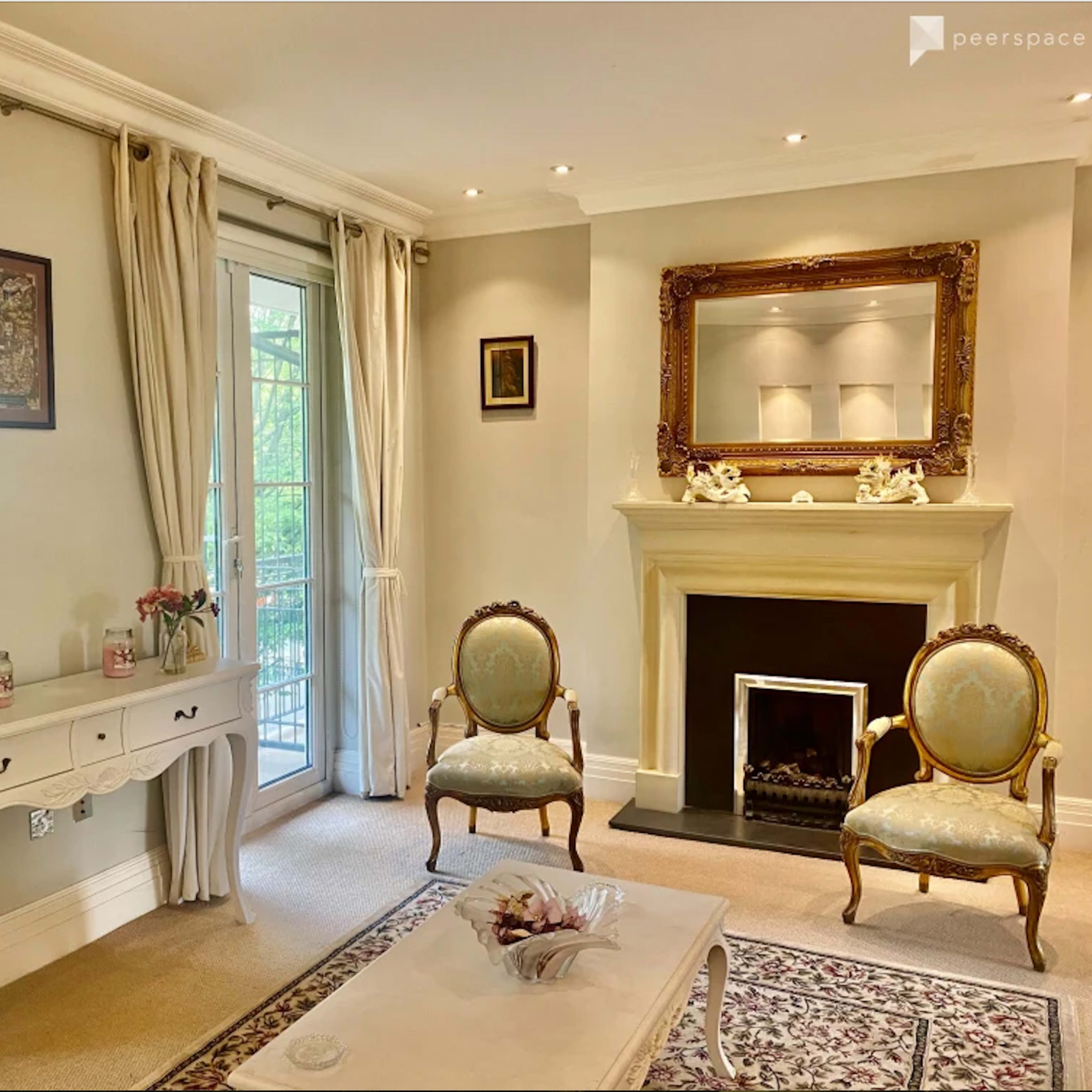 A elegantly decorated living room featuring a fireplace, a decorative mirror, two antique chairs, and a console table with floral decor.