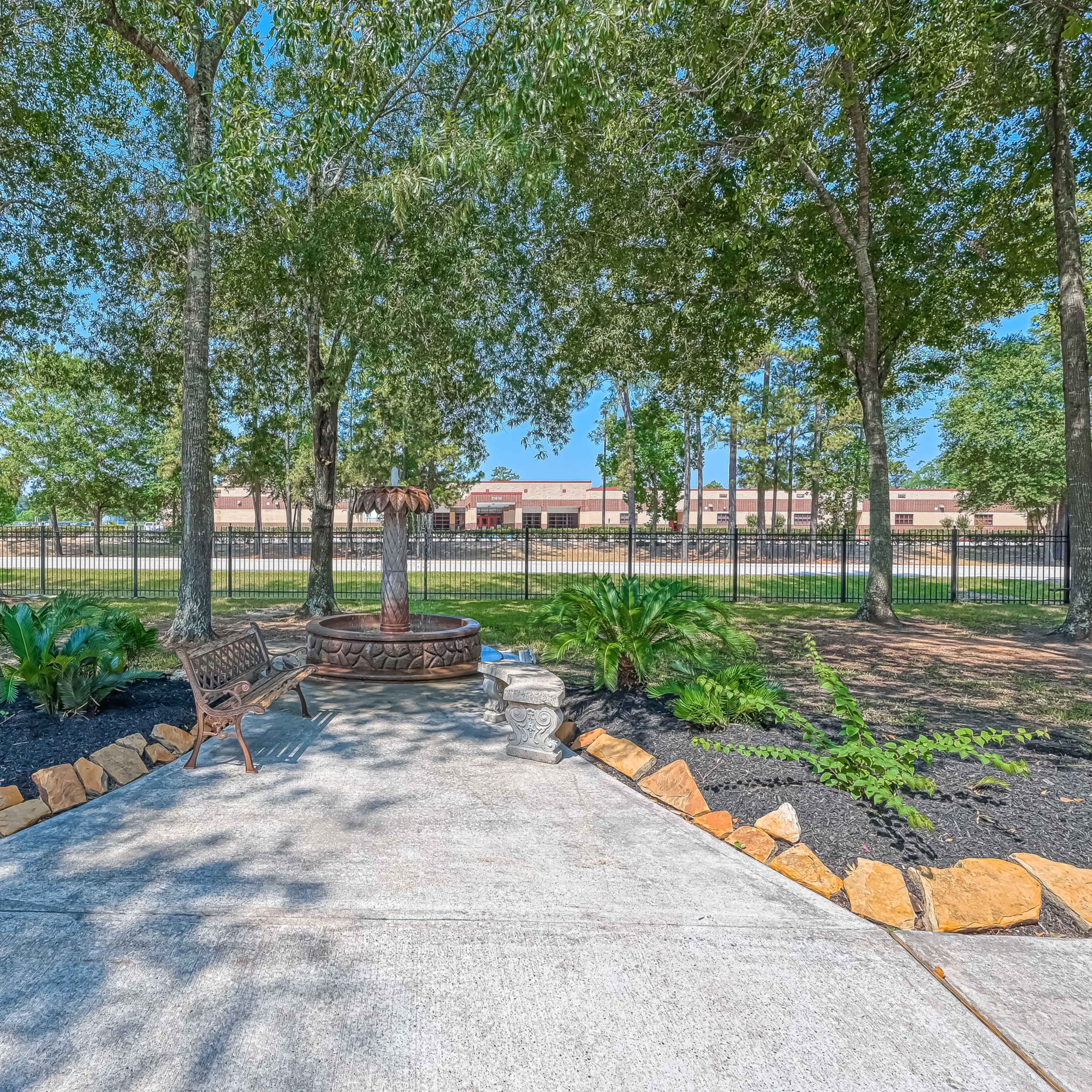 A landscaped area features a stone-edged pathway leading to a fountain, surrounded by trees and shrubs with a view of a fence in the background.