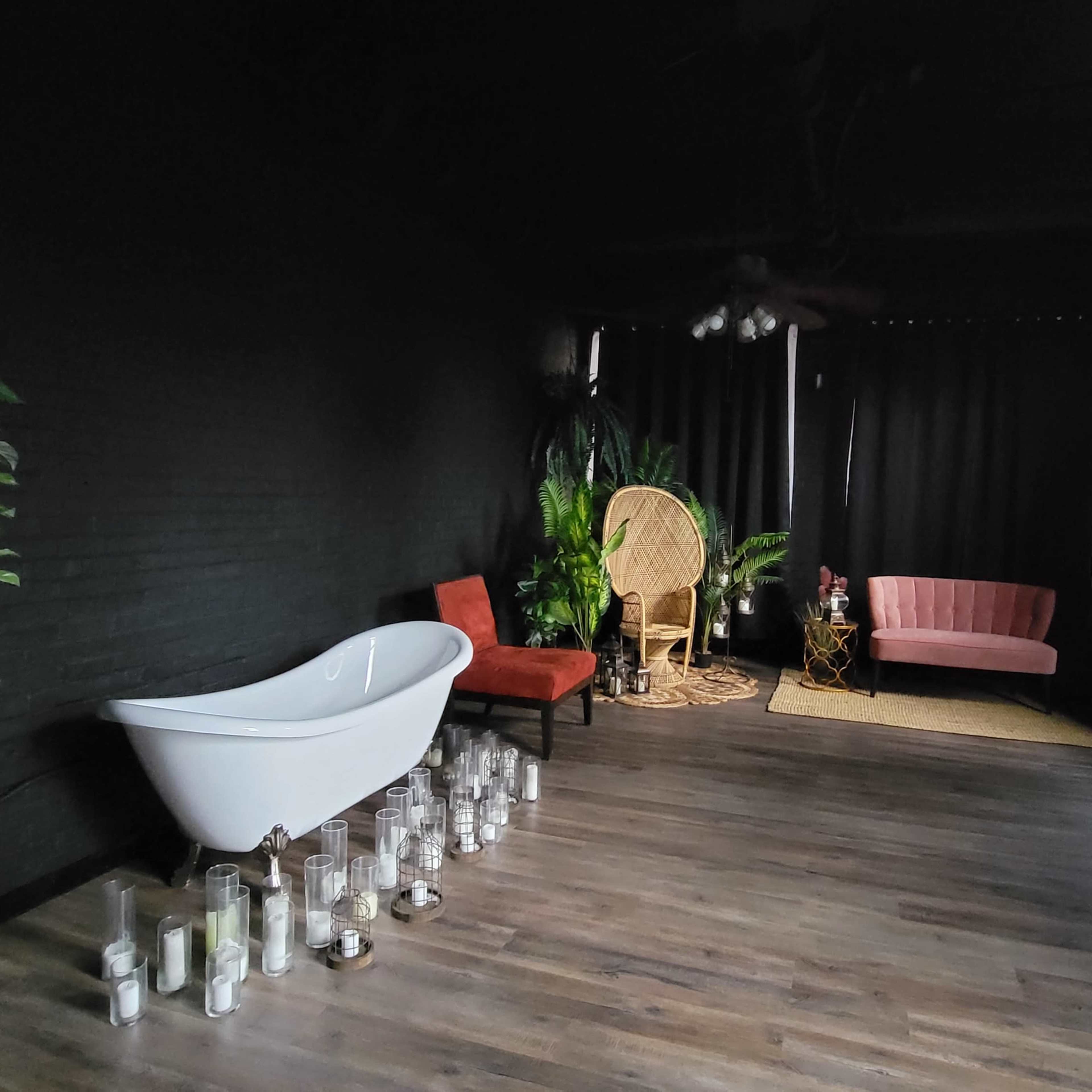 A dimly lit room with a freestanding white bathtub, two chairs in contrasting colors, a large plant, and several candles arranged on the floor.