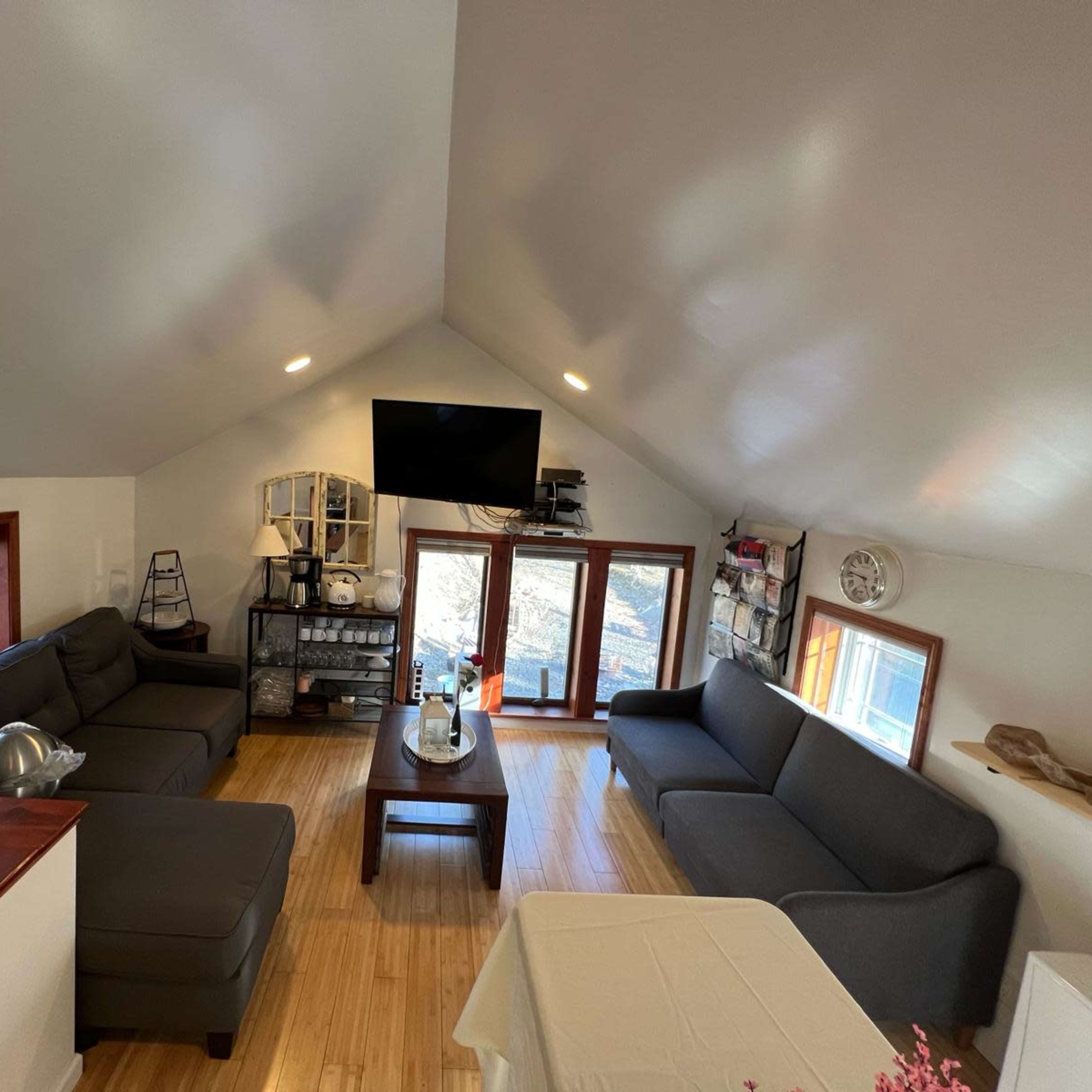 A bright, spacious living room with two gray sofas, a coffee table, a large TV mounted on the wall, and large windows providing natural light.
