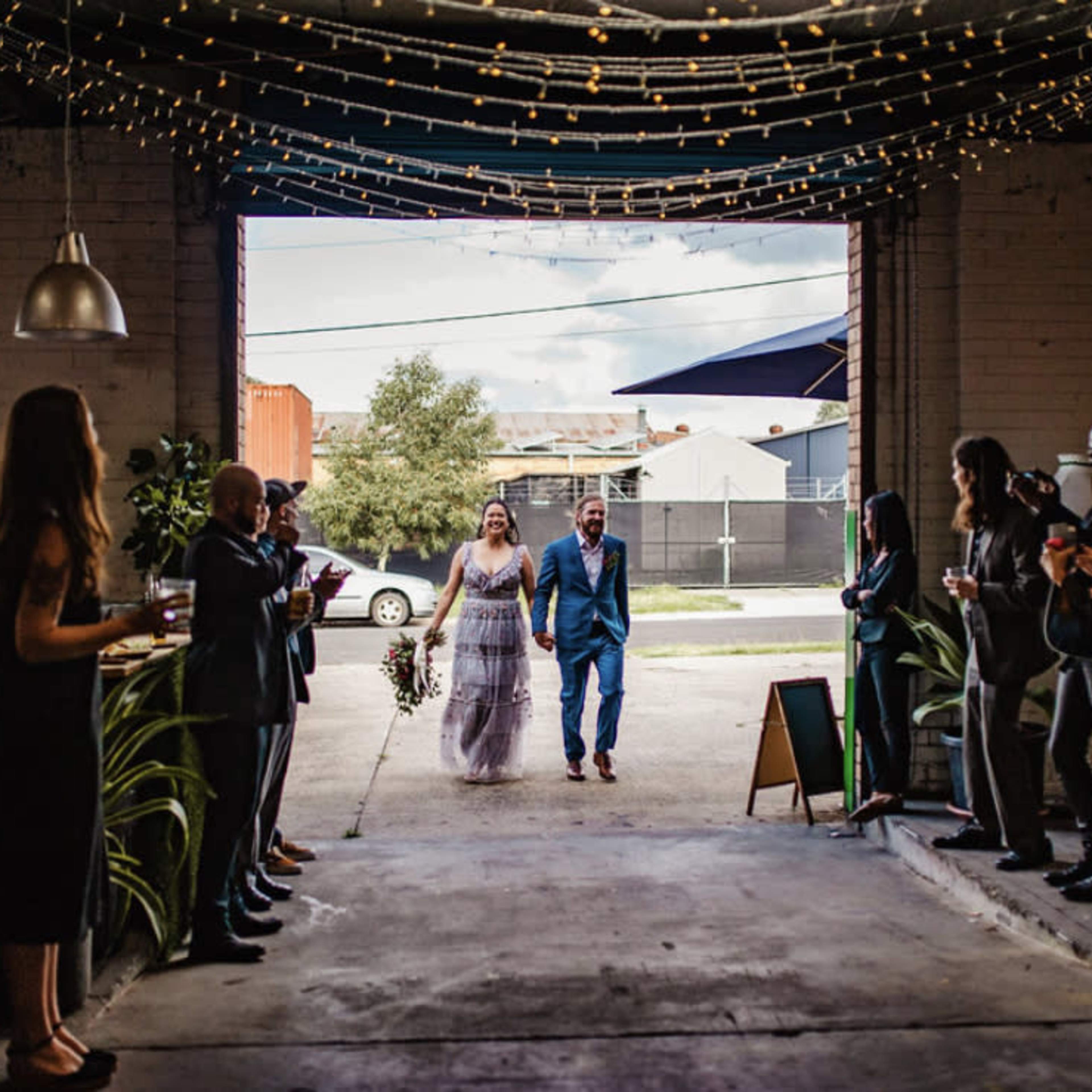 A bride and groom walk hand in hand through a doorway, greeted by guests clapping on either side in an industrial venue.