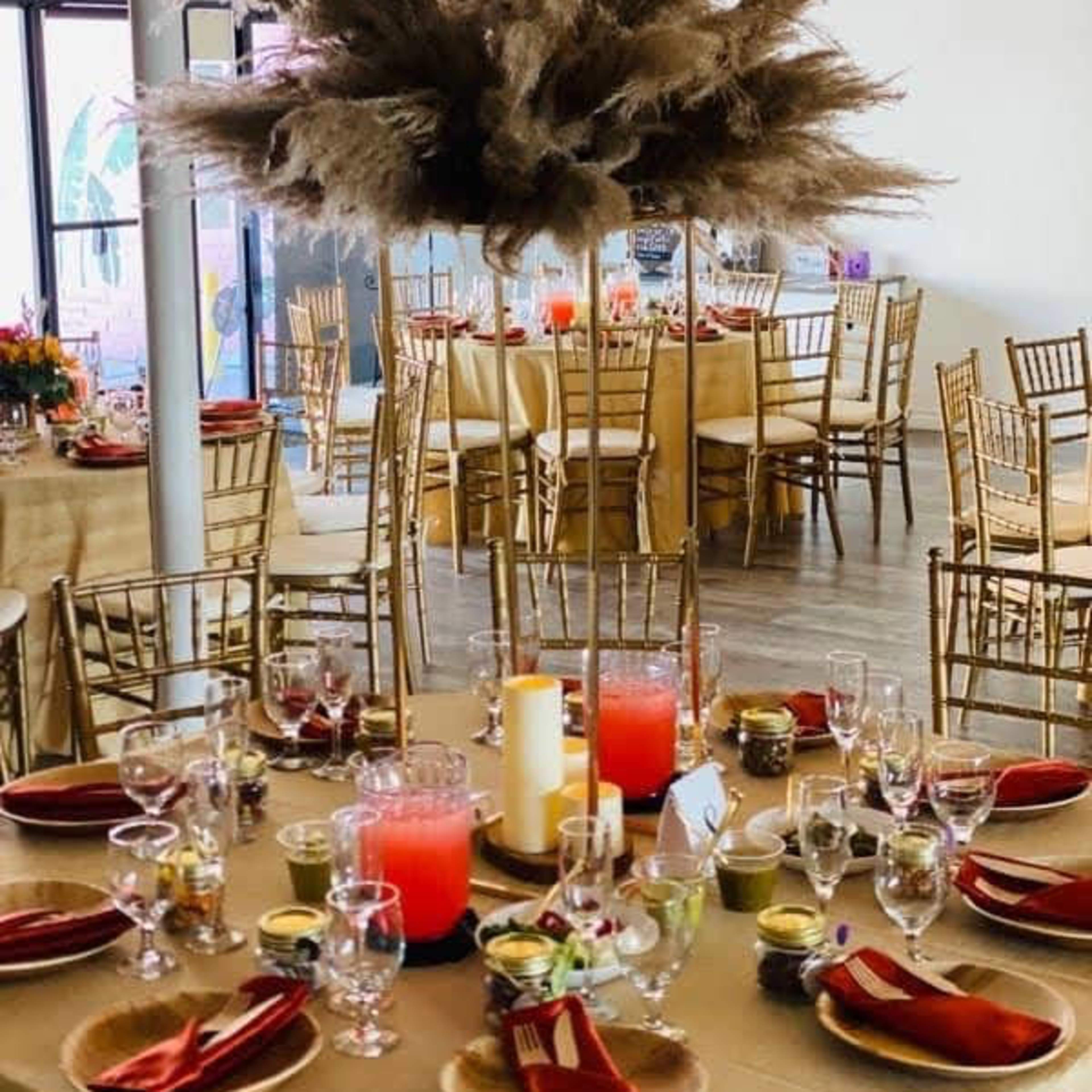 A beautifully arranged dining setup features golden tables adorned with tall centerpieces made of pampas grass, surrounded by elegant chairs and tableware in a well-lit venue.