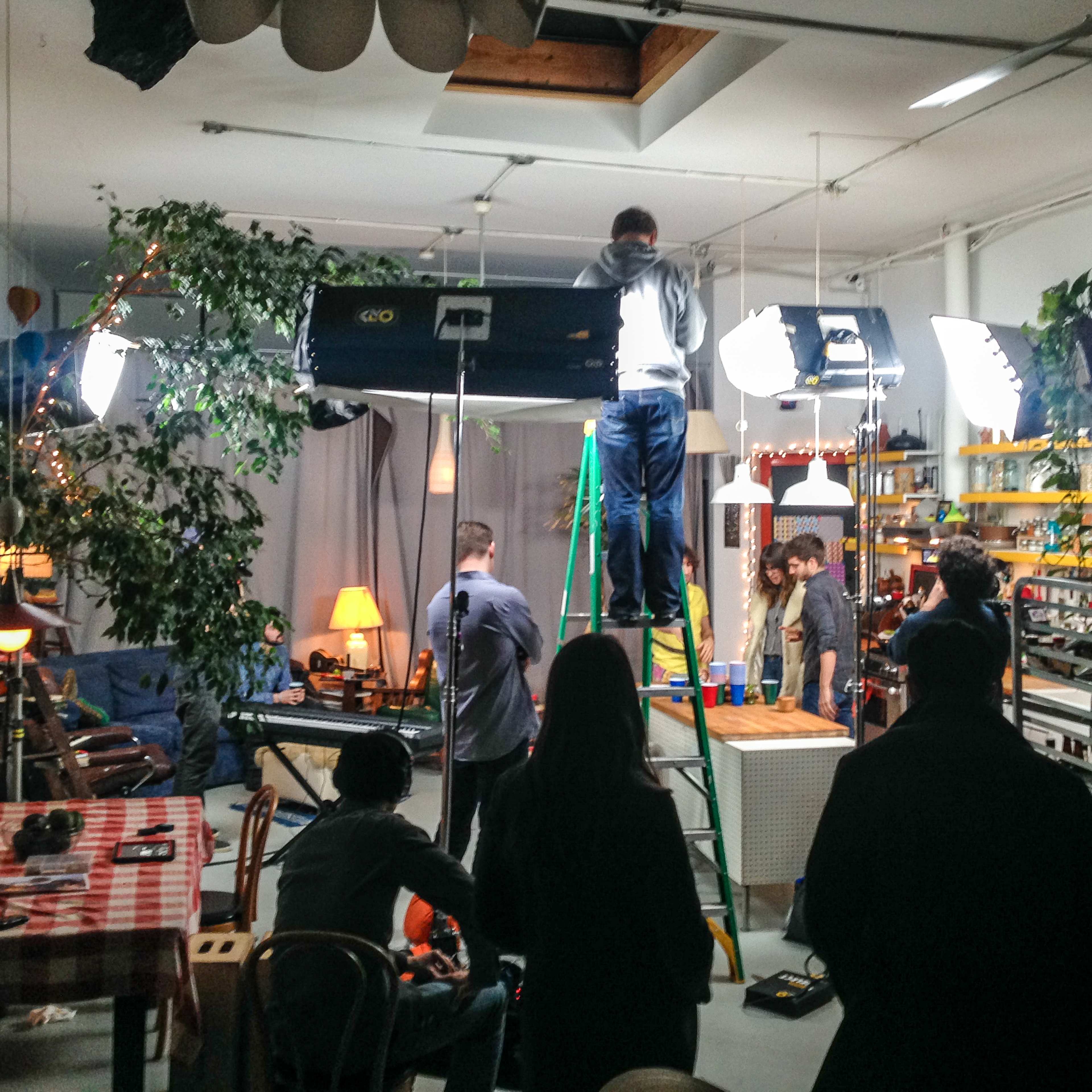 A film crew sets up lighting equipment in a cozy indoor set while others prepare for a shoot.