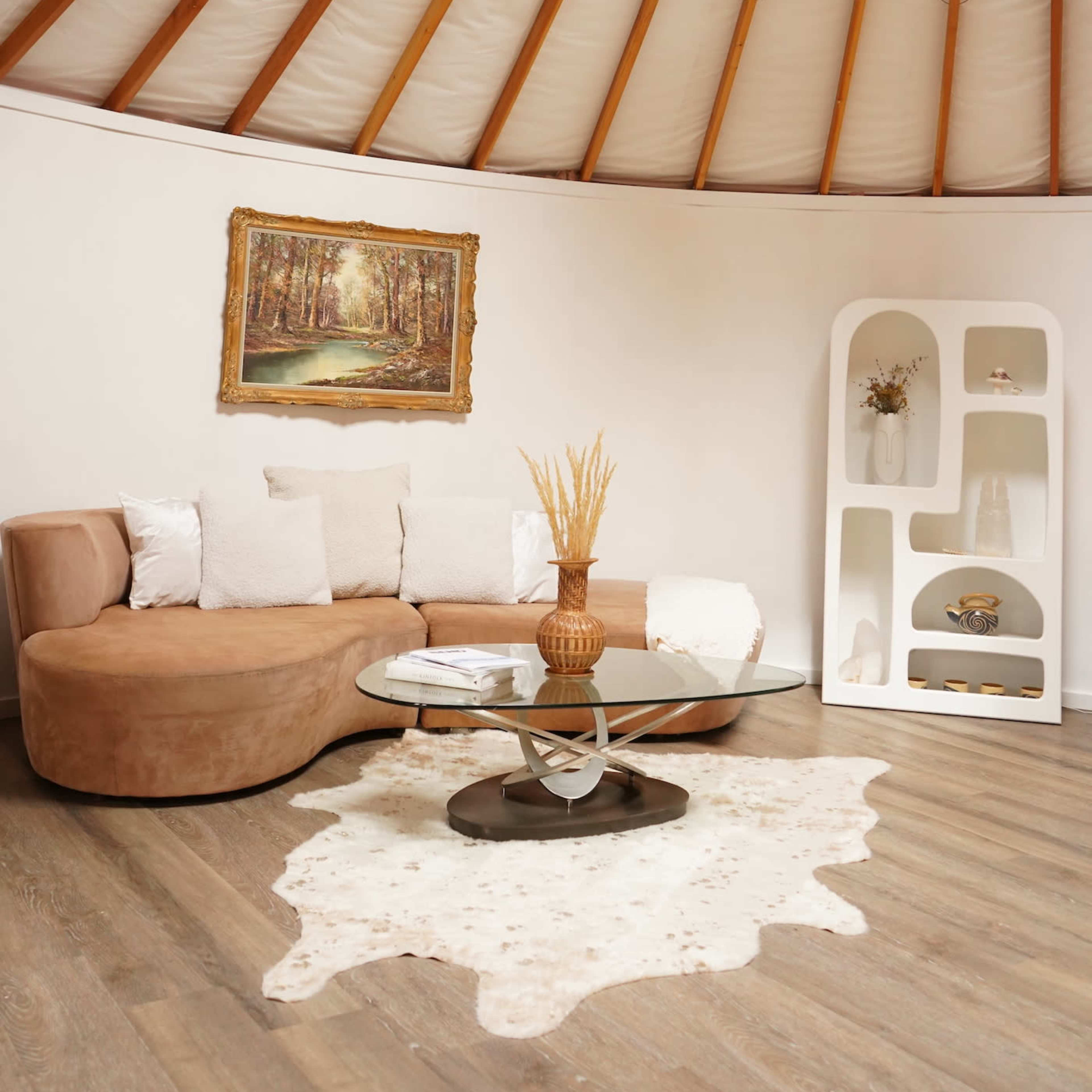 A modern circular room with wooden beams, featuring a curved brown sofa, a round coffee table, a decorative vase, and a framed painting of a forest.
