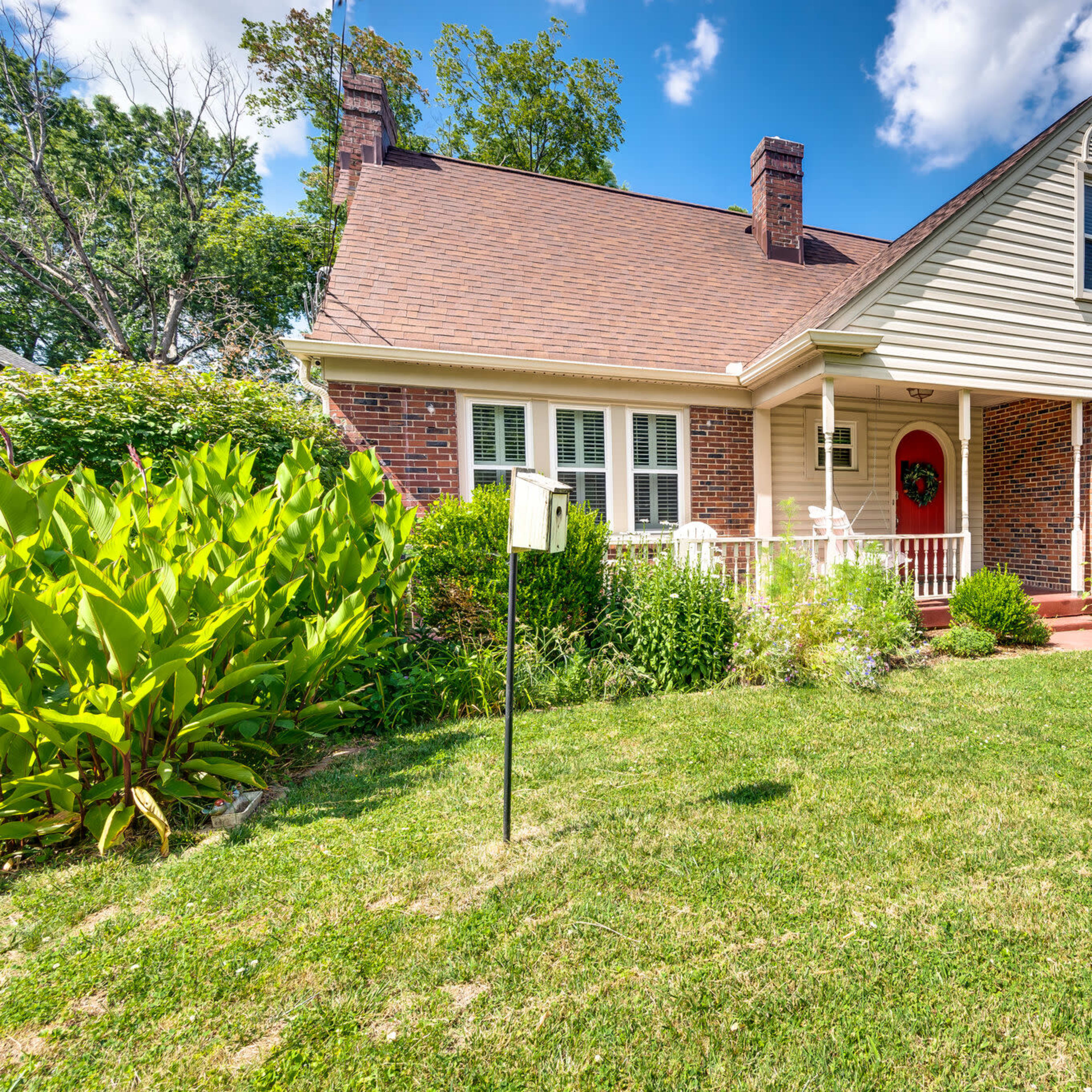 A brick house with a front porch and a red door is surrounded by green shrubs and grass under a blue sky.