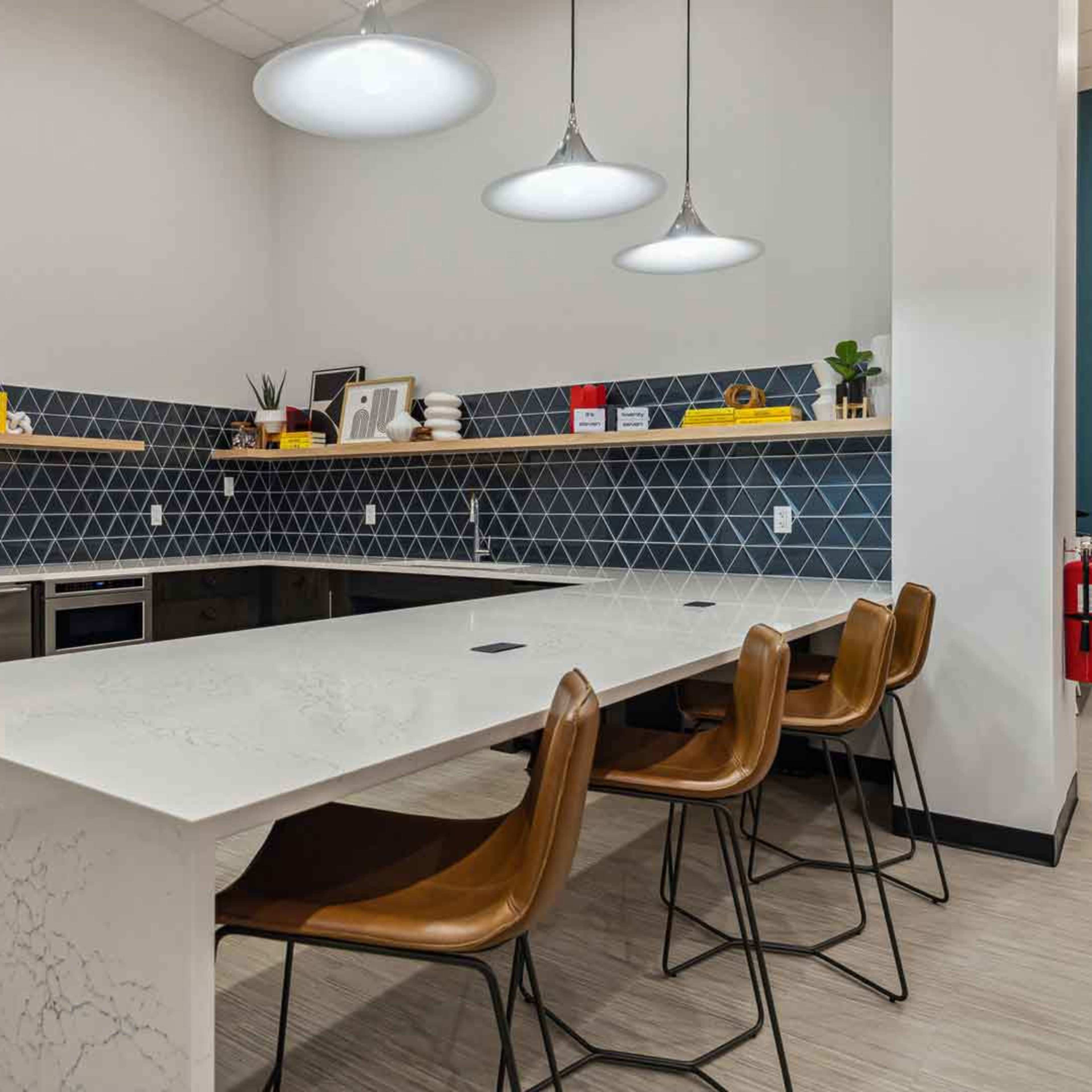 A modern kitchen with a large white countertop, four brown chairs, and geometric tile backsplash featuring decorative items on shelves.