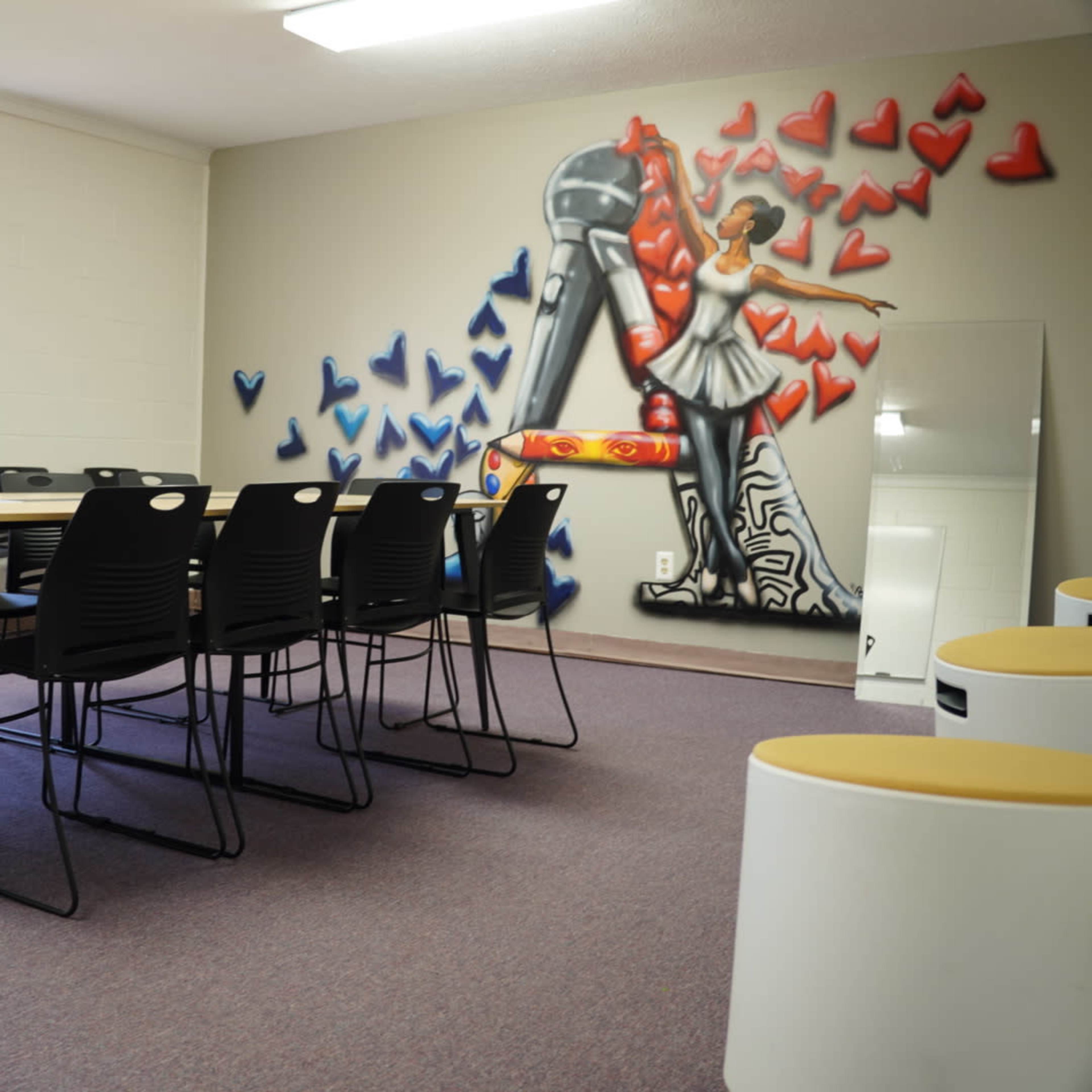 A room with a table surrounded by black chairs and a mural of a figure in front of a large red and blue heart design on the wall.
