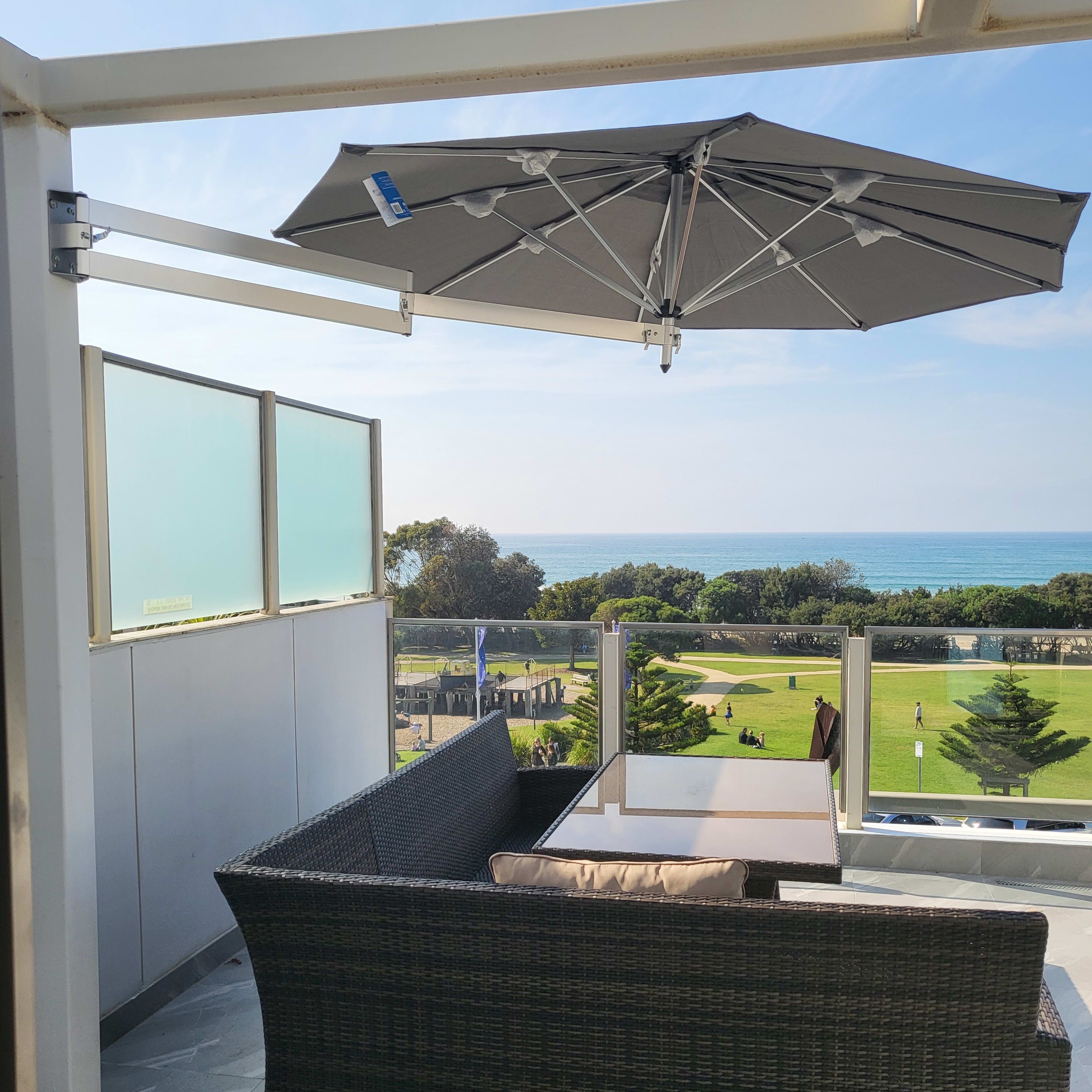 A balcony features a patio set with a table and a large umbrella, overlooking a grassy area and the ocean in the distance.