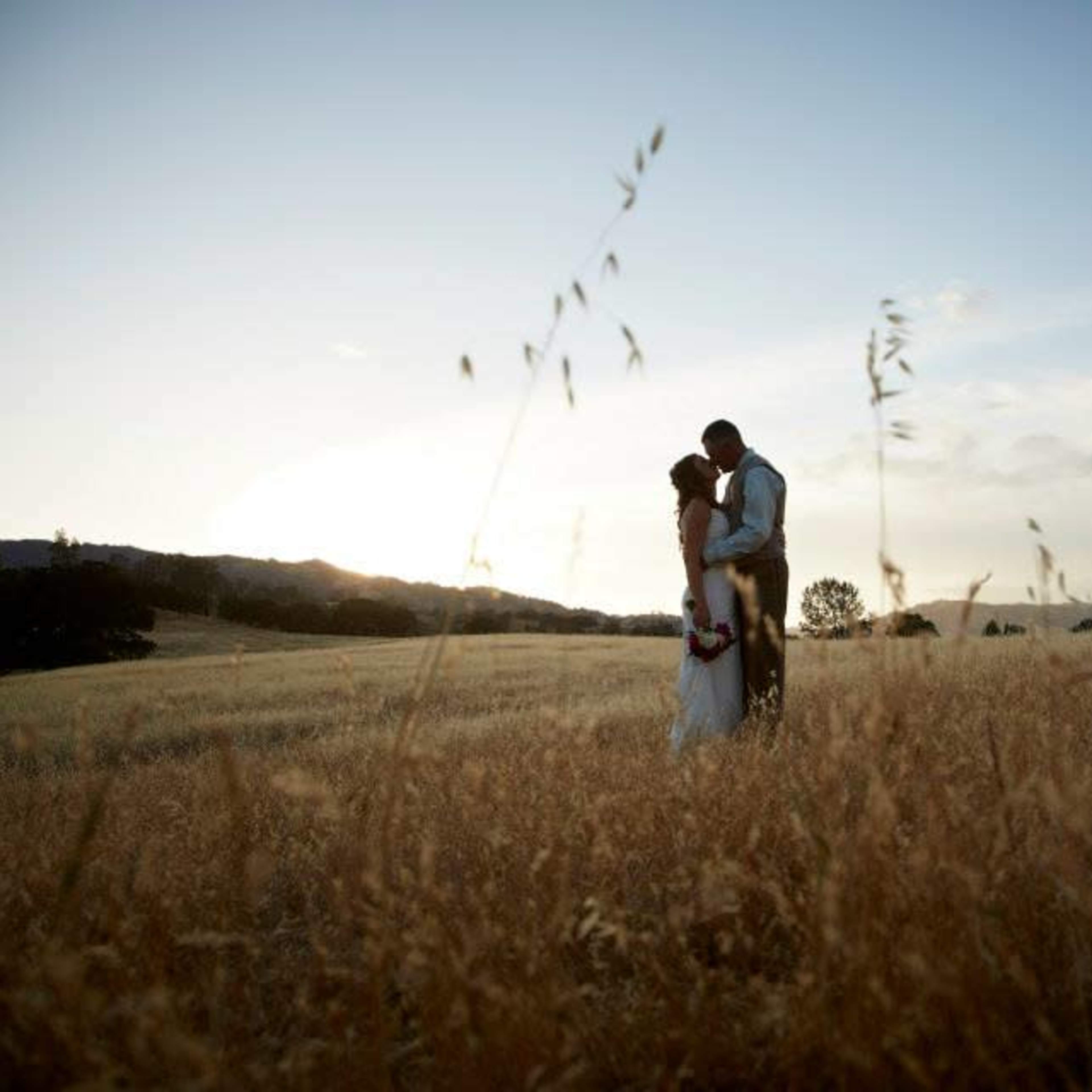 A couple embraces in a grassy field at sunset, surrounded by tall grass and distant trees.