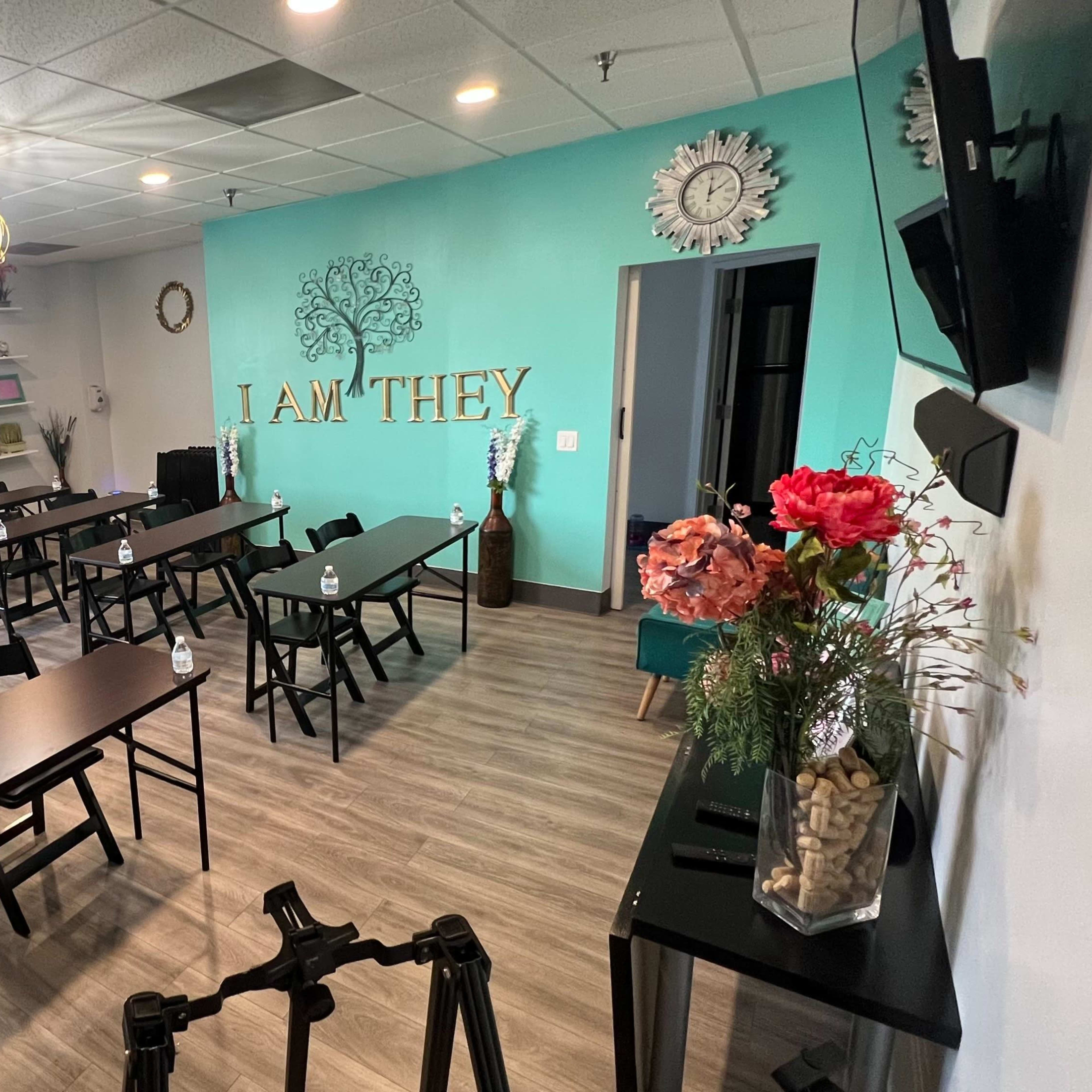 A classroom or meeting space with multiple rows of black tables and chairs, a turquoise wall, and decorative elements including flowers and wall art.