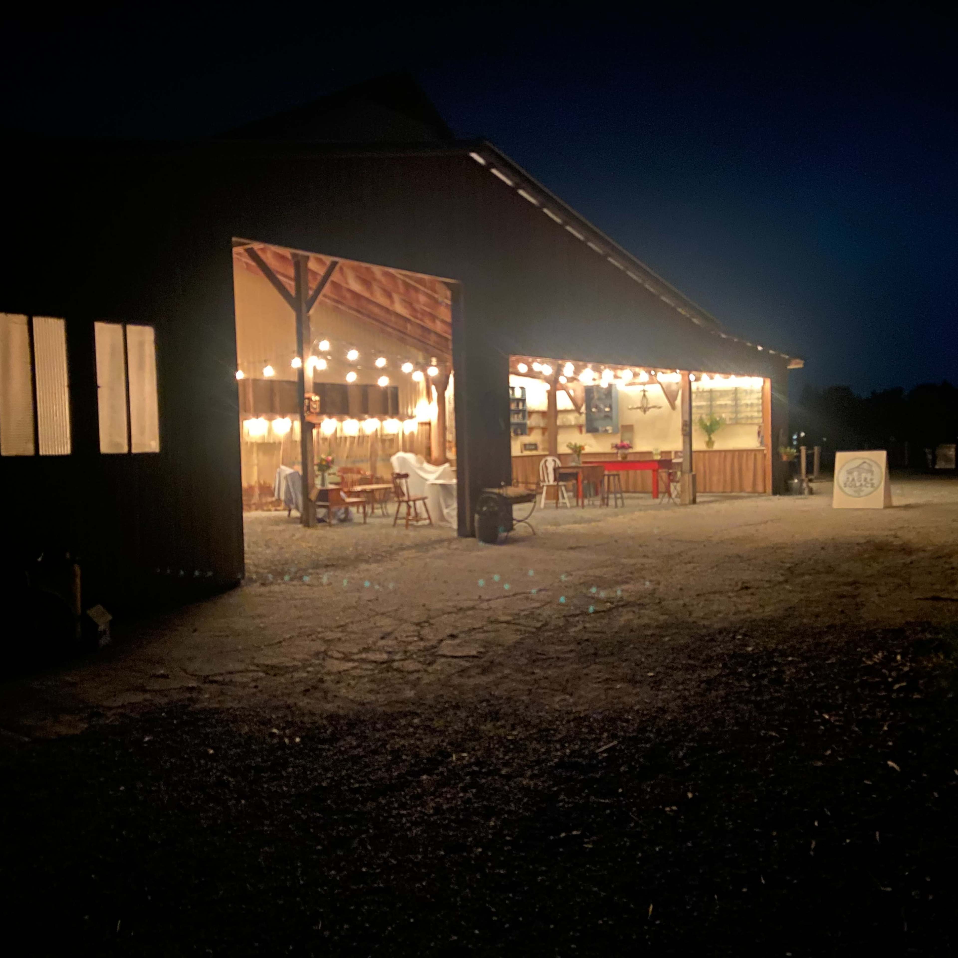 A barn-like structure is illuminated at night, revealing tables and chairs arranged inside under string lights.