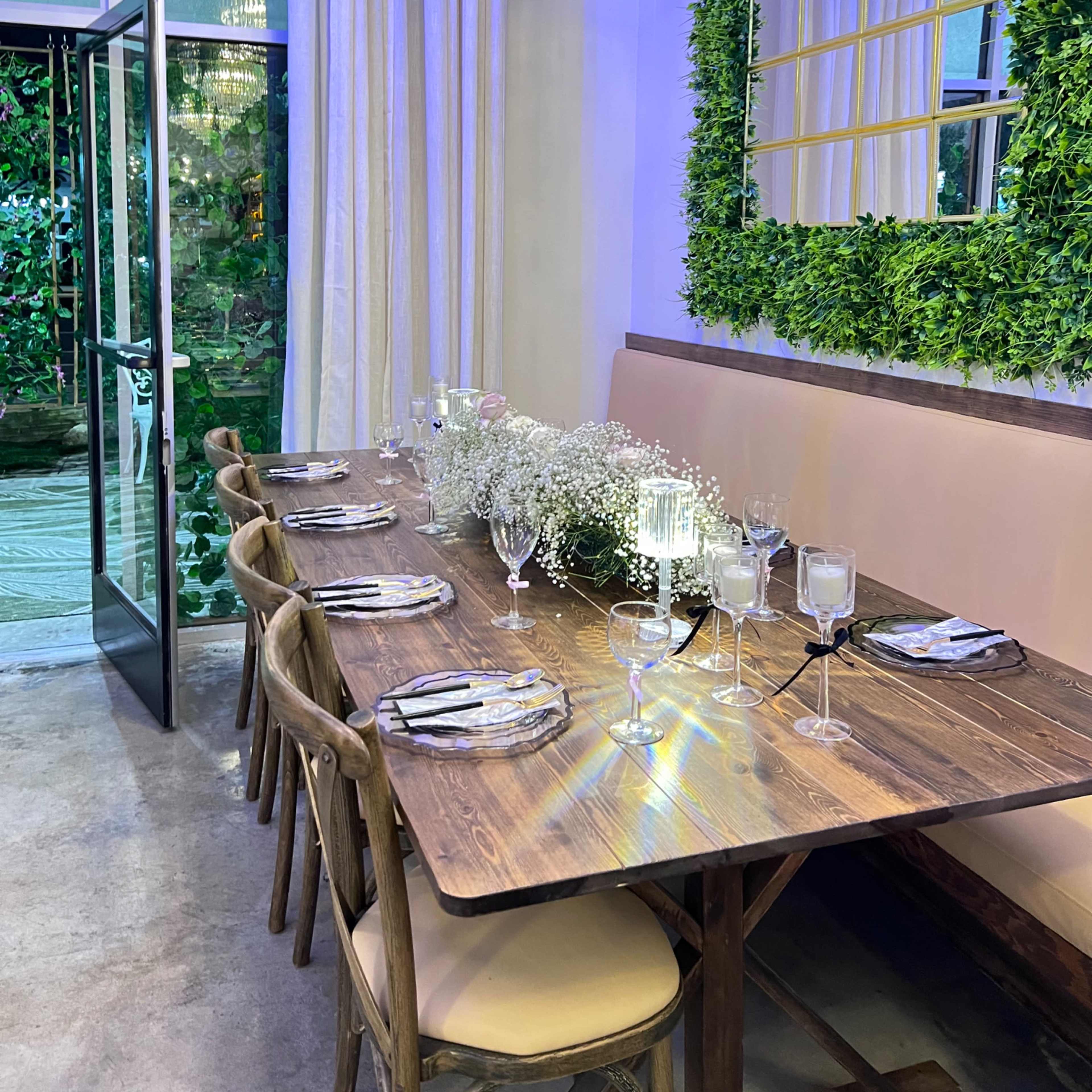 A dining table is set with elegant tableware and a floral centerpiece in a room featuring large windows and greenery accents on the walls.