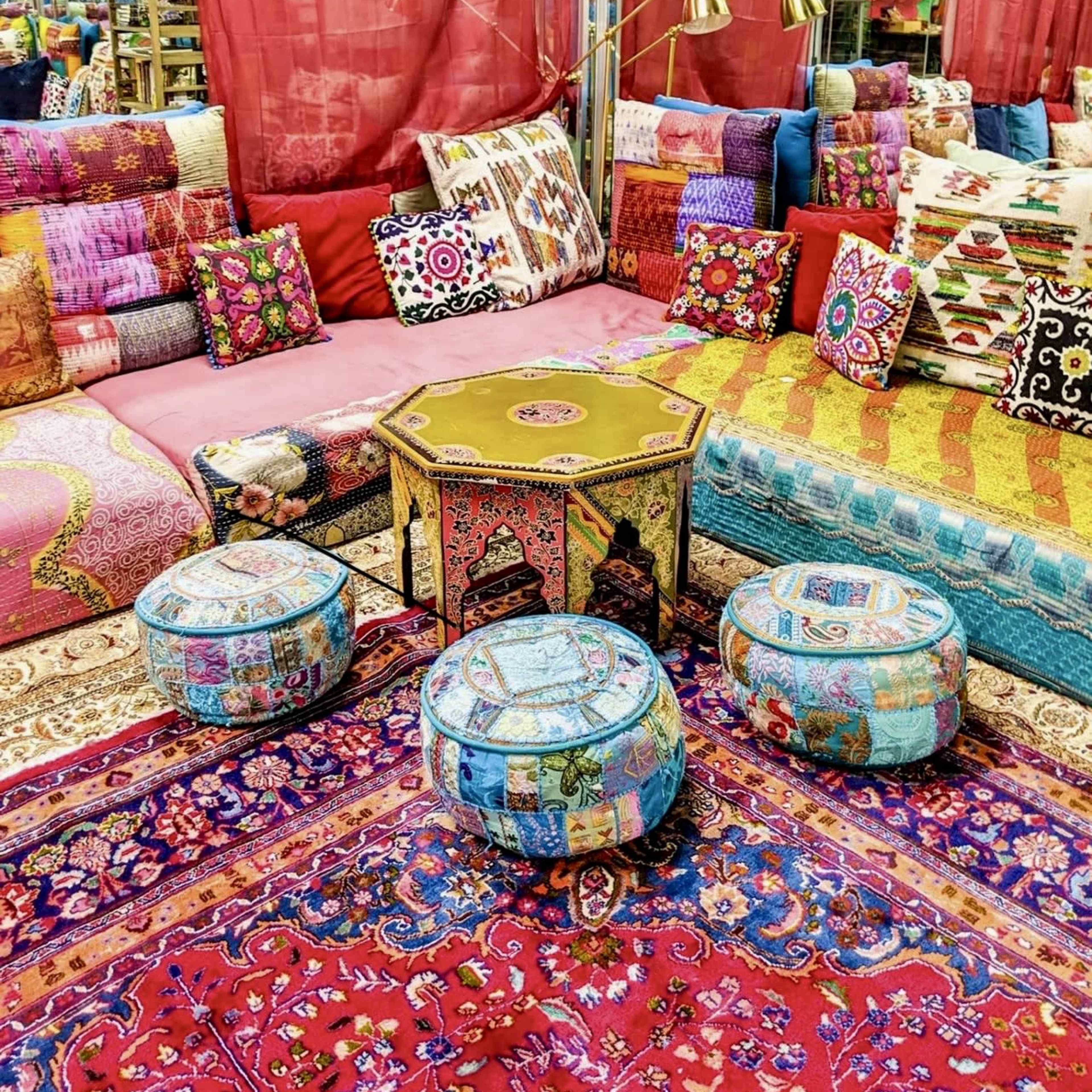 The scene features a colorful arrangement of cushions, pillows, and poufs surrounding a decorative table in an indoor space with vibrant textiles and drapes.