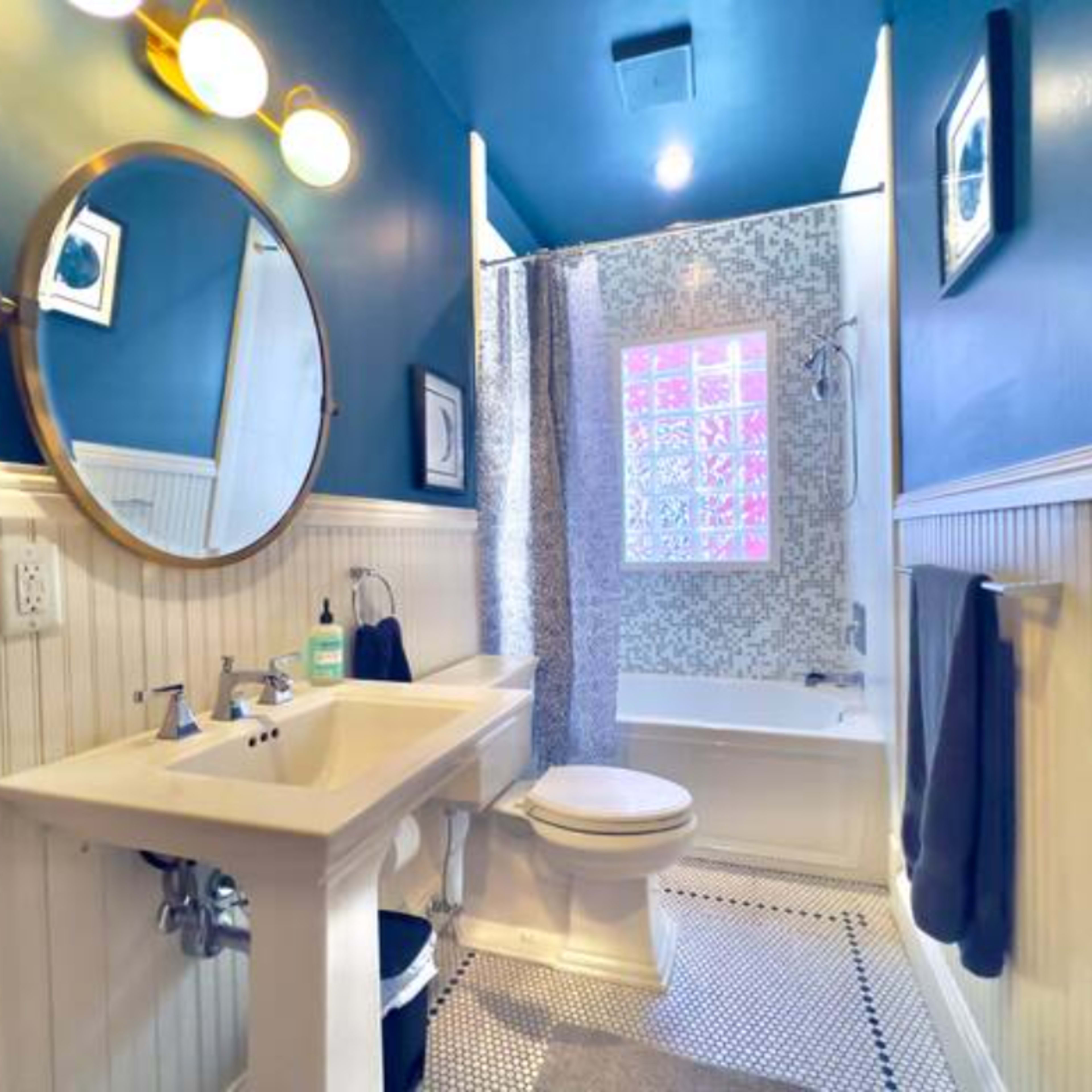 A small bathroom with a circular mirror, a white sink, a toilet, a shower area with a patterned curtain, and blue walls.