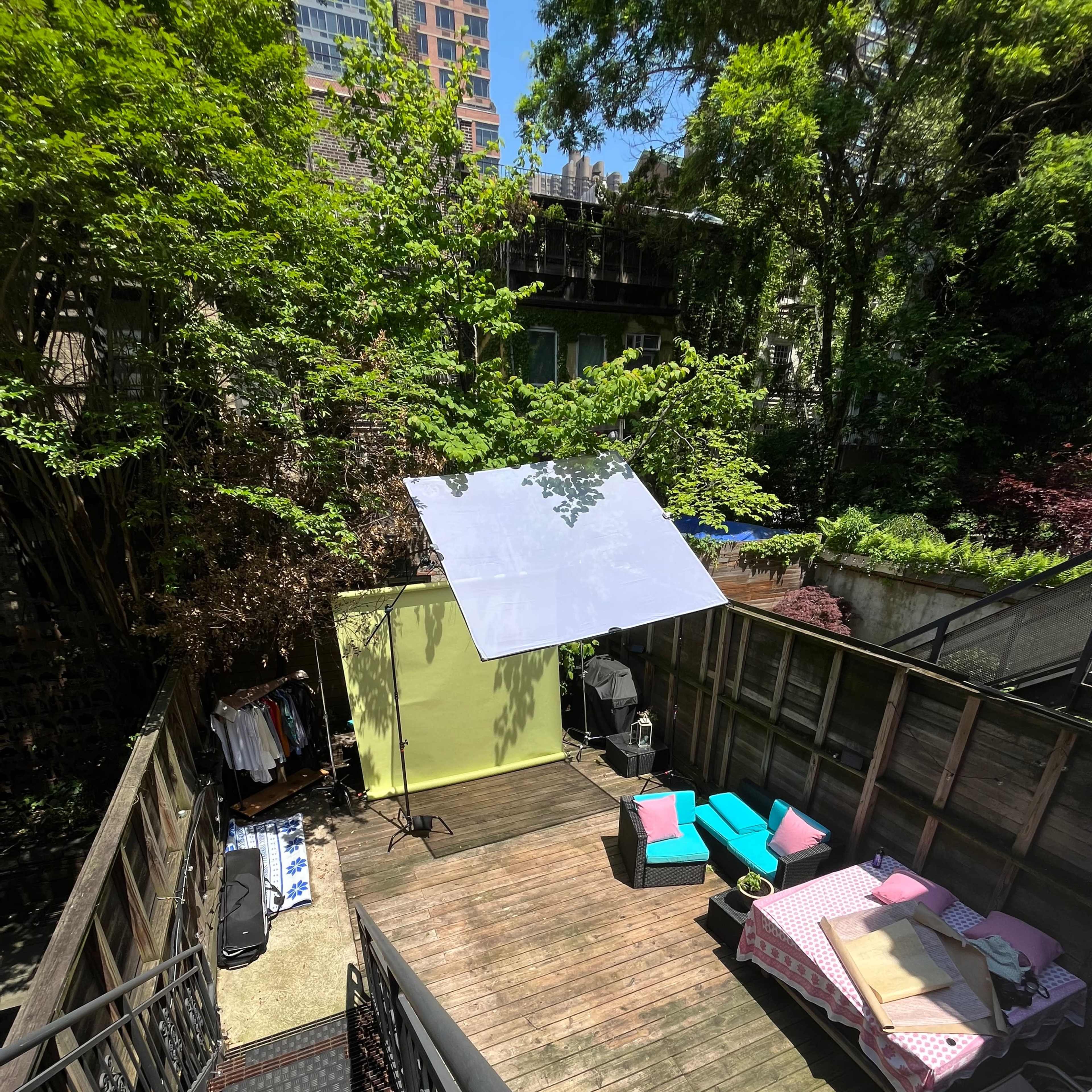 A wooden deck area with a seating arrangement, a large white backdrop, and a few clothes hanging in a lush, green environment.