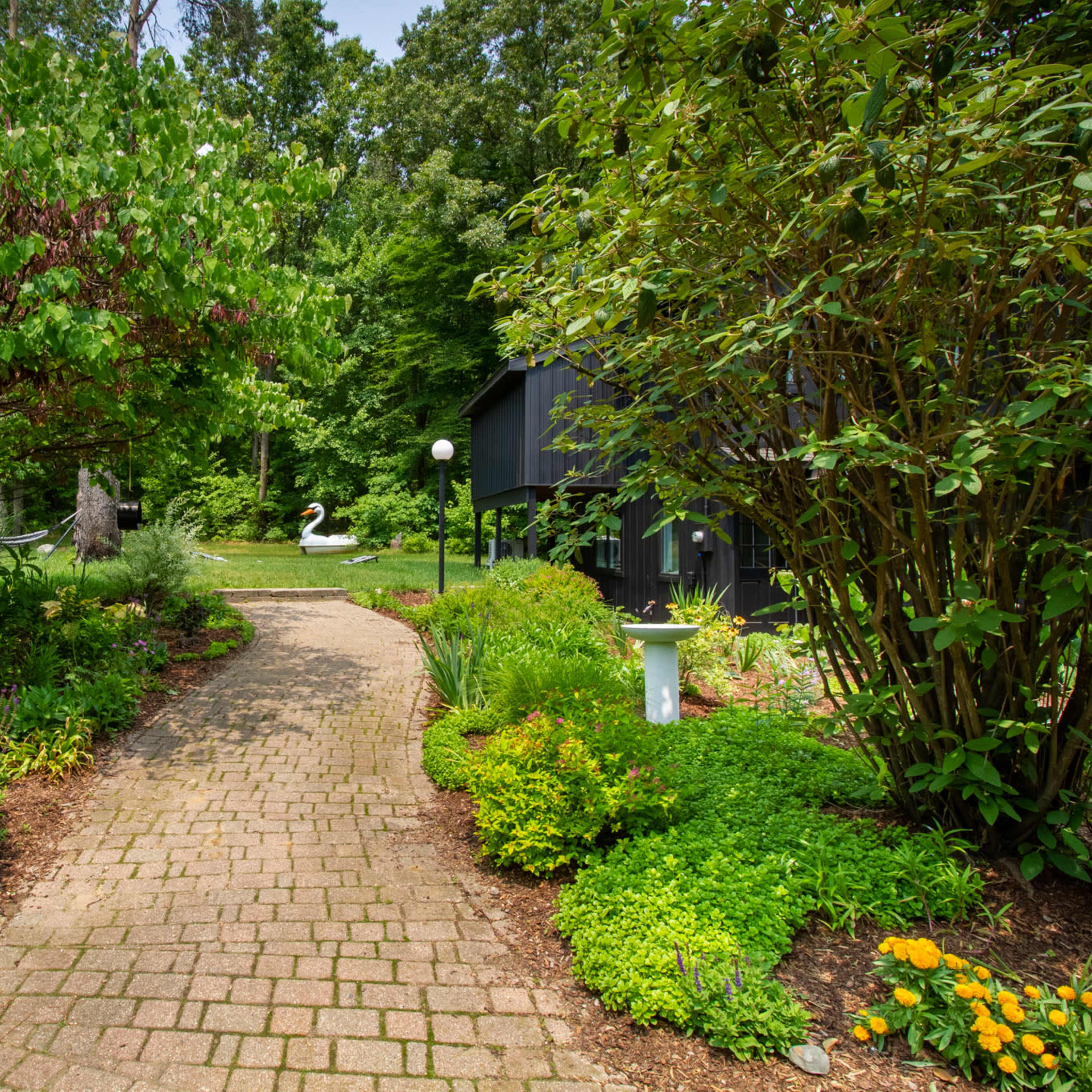 A brick pathway winds through lush greenery, leading to a pond with a swan in the background.