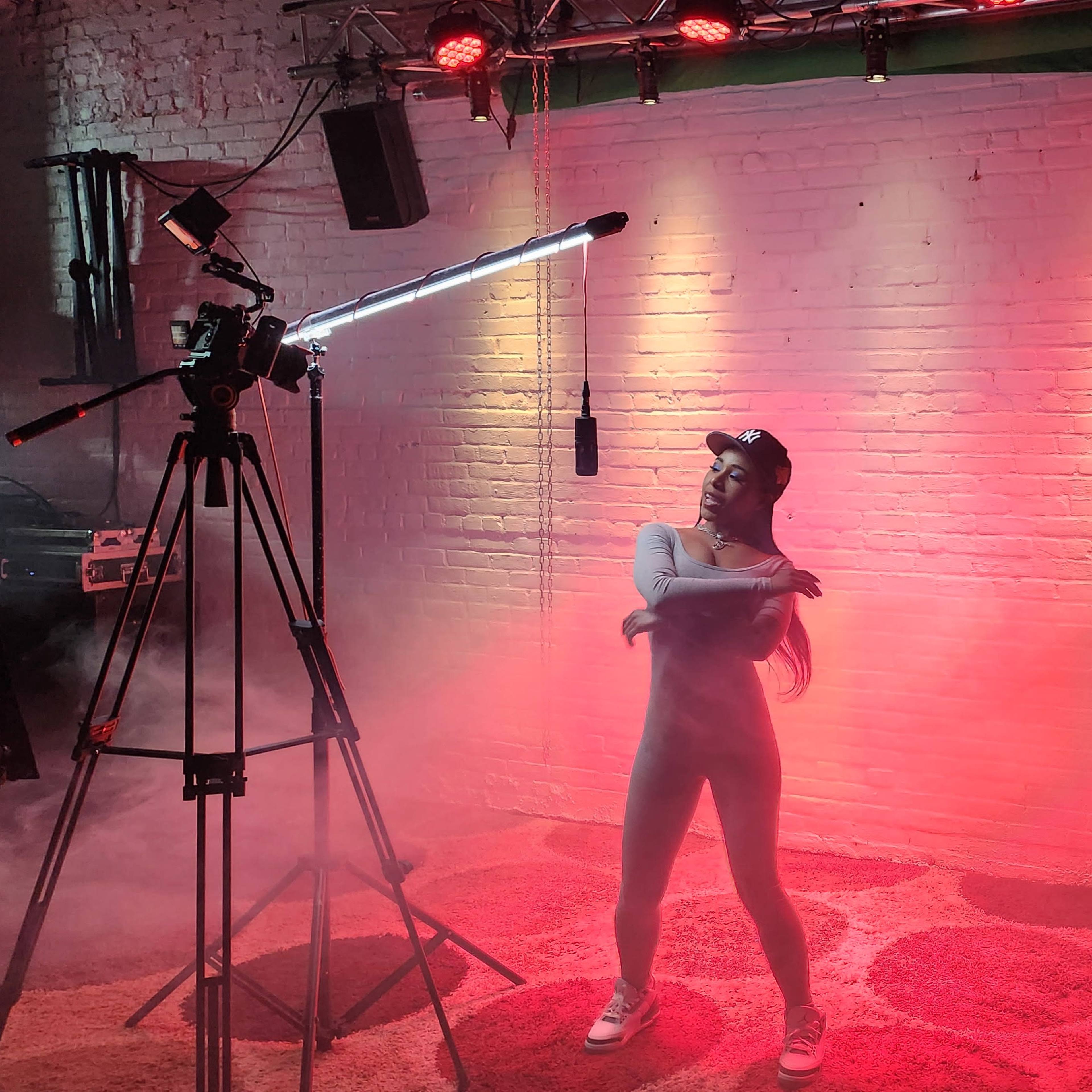 A performer poses in front of a camera setup with colorful lights and fog in a studio setting.