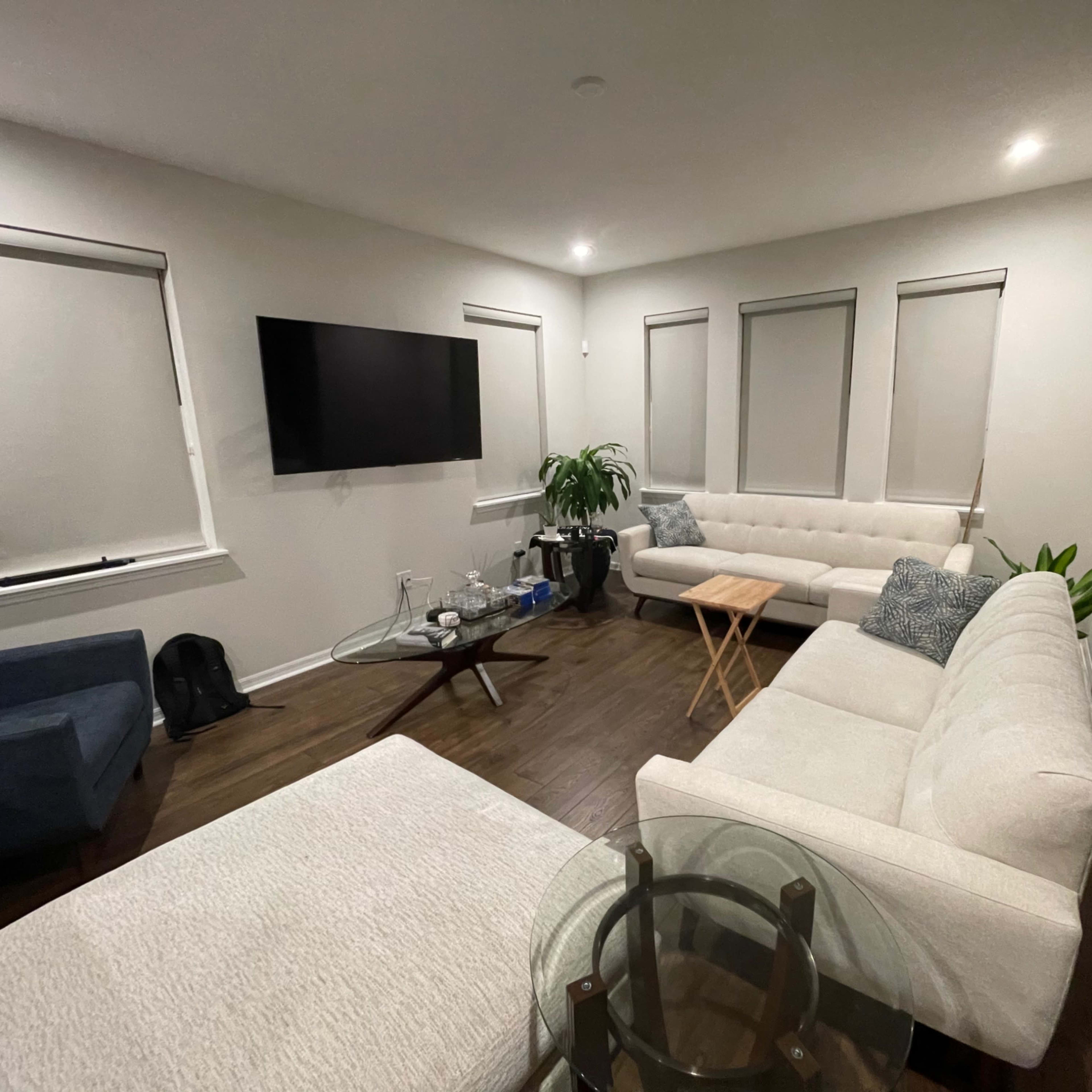 A modern living room with a large television mounted on the wall, a light-colored sectional sofa, a blue armchair, and a small wooden table.