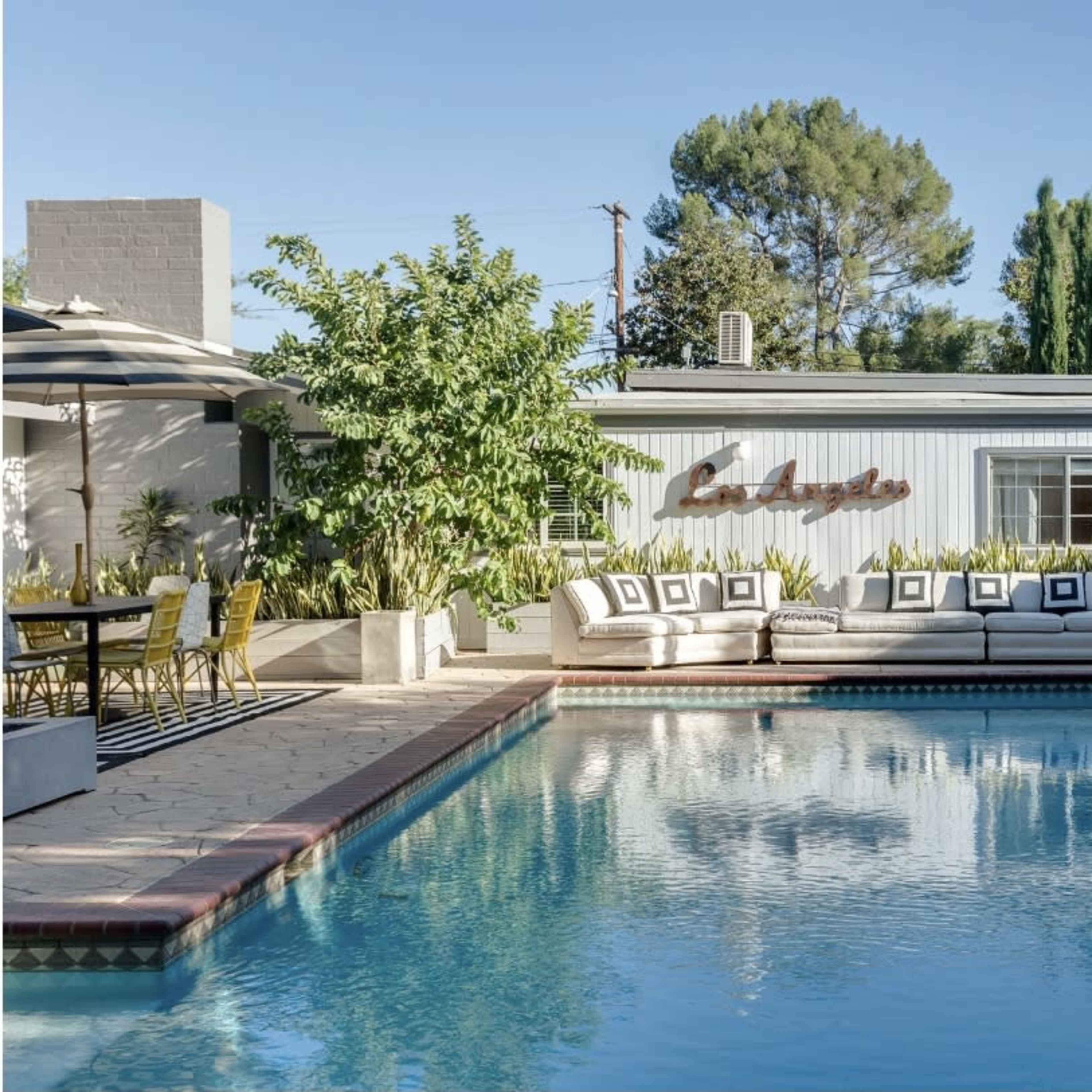 A swimming pool reflects a seating area and a house with large windows, surrounded by trees and landscaped gardens.
