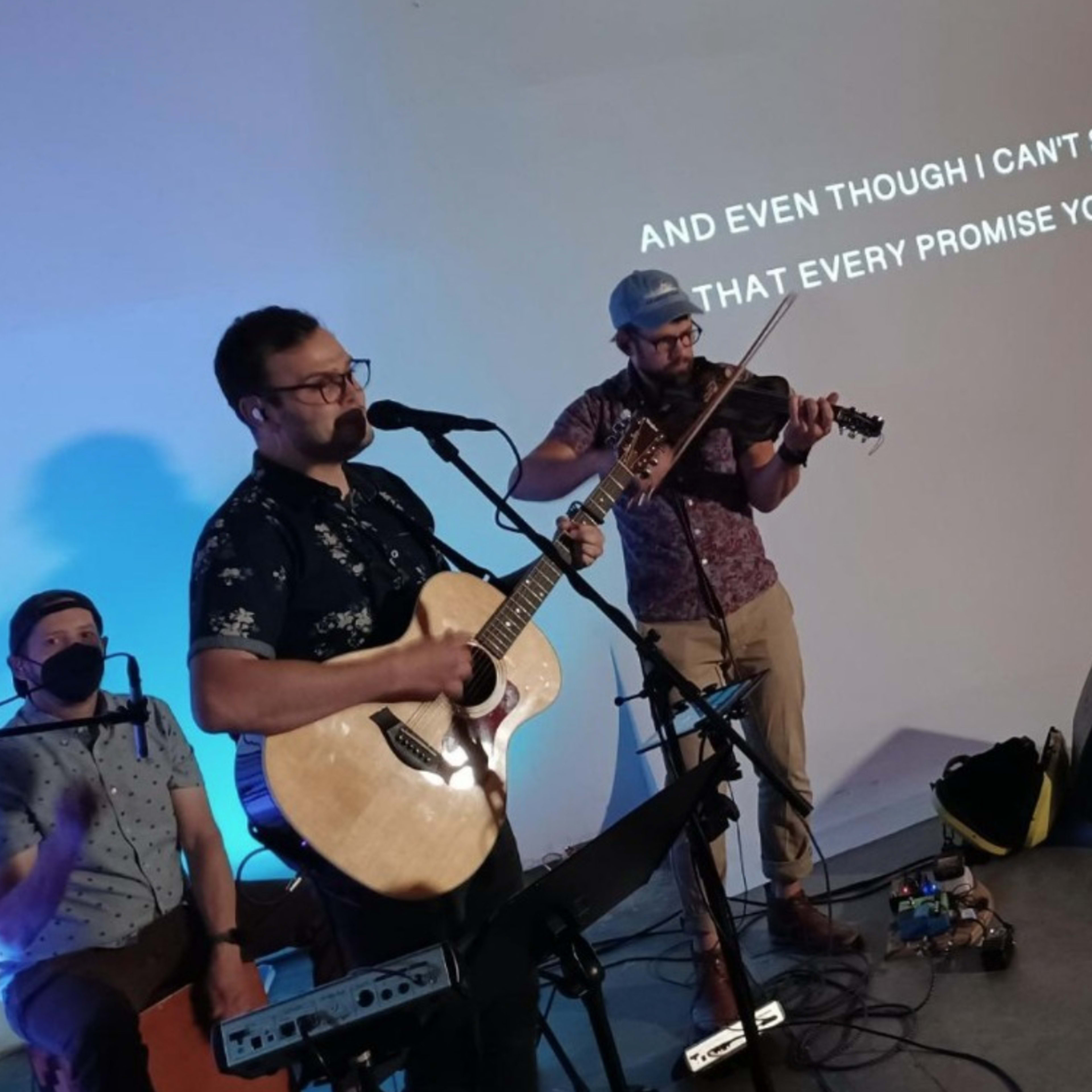 Two musicians perform on stage, with one playing guitar and singing while the other plays the violin, accompanied by lyrics displayed on a screen behind them.
