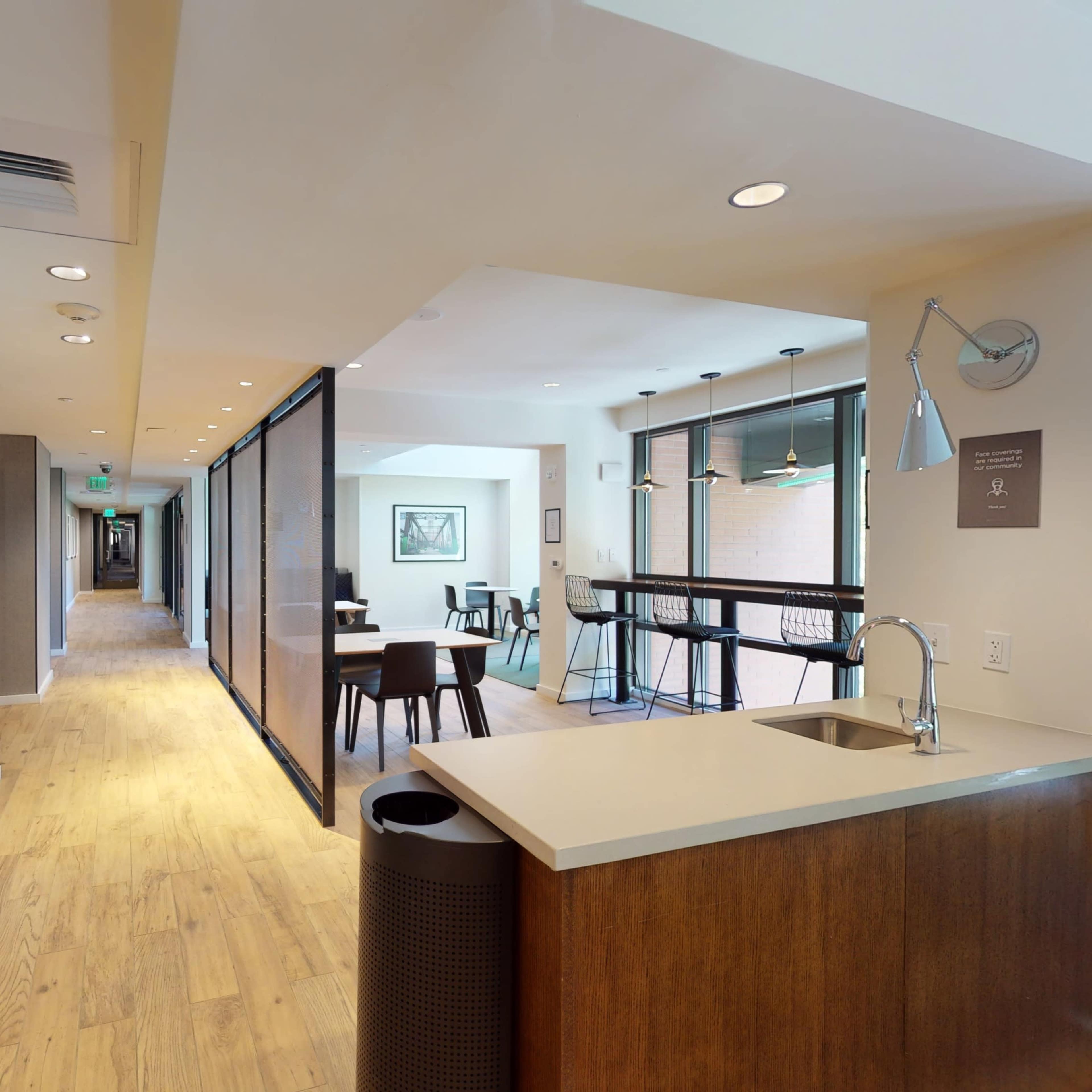 A modern hallway that connects a kitchen area to a seating area, featuring a mix of wooden flooring and light-colored walls, with glass panel dividers.
