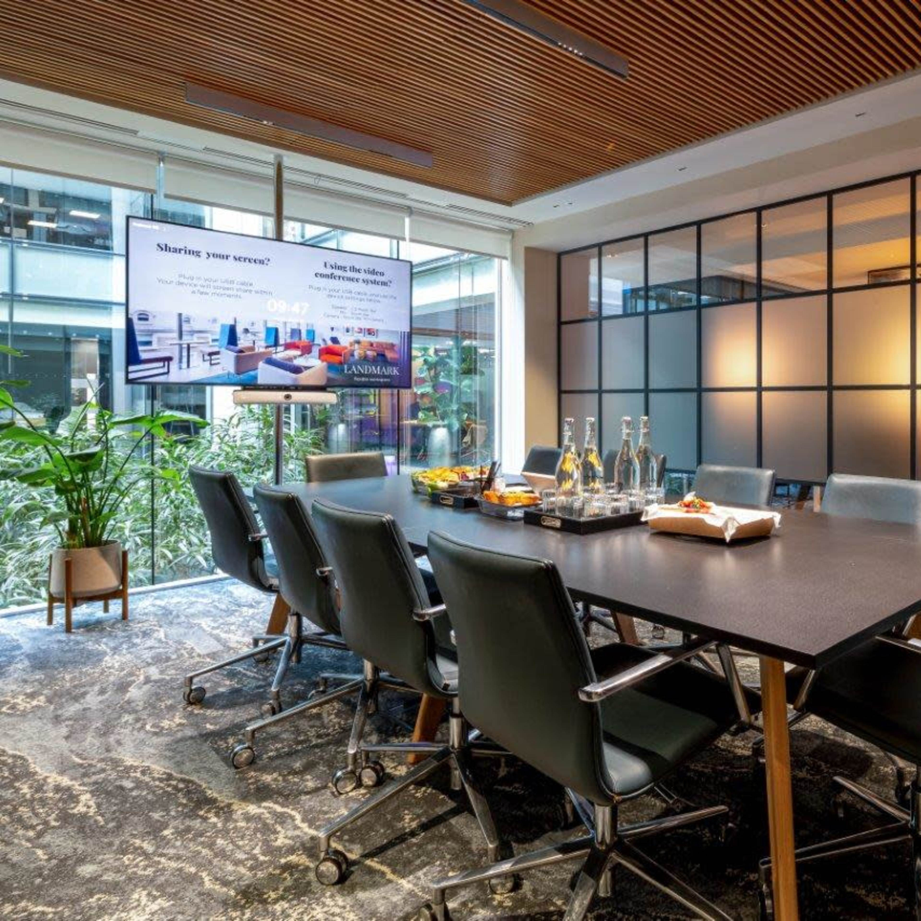 A modern conference room with a large table surrounded by office chairs, a screen displaying a presentation, and glass walls providing a view of an indoor plant and natural light.