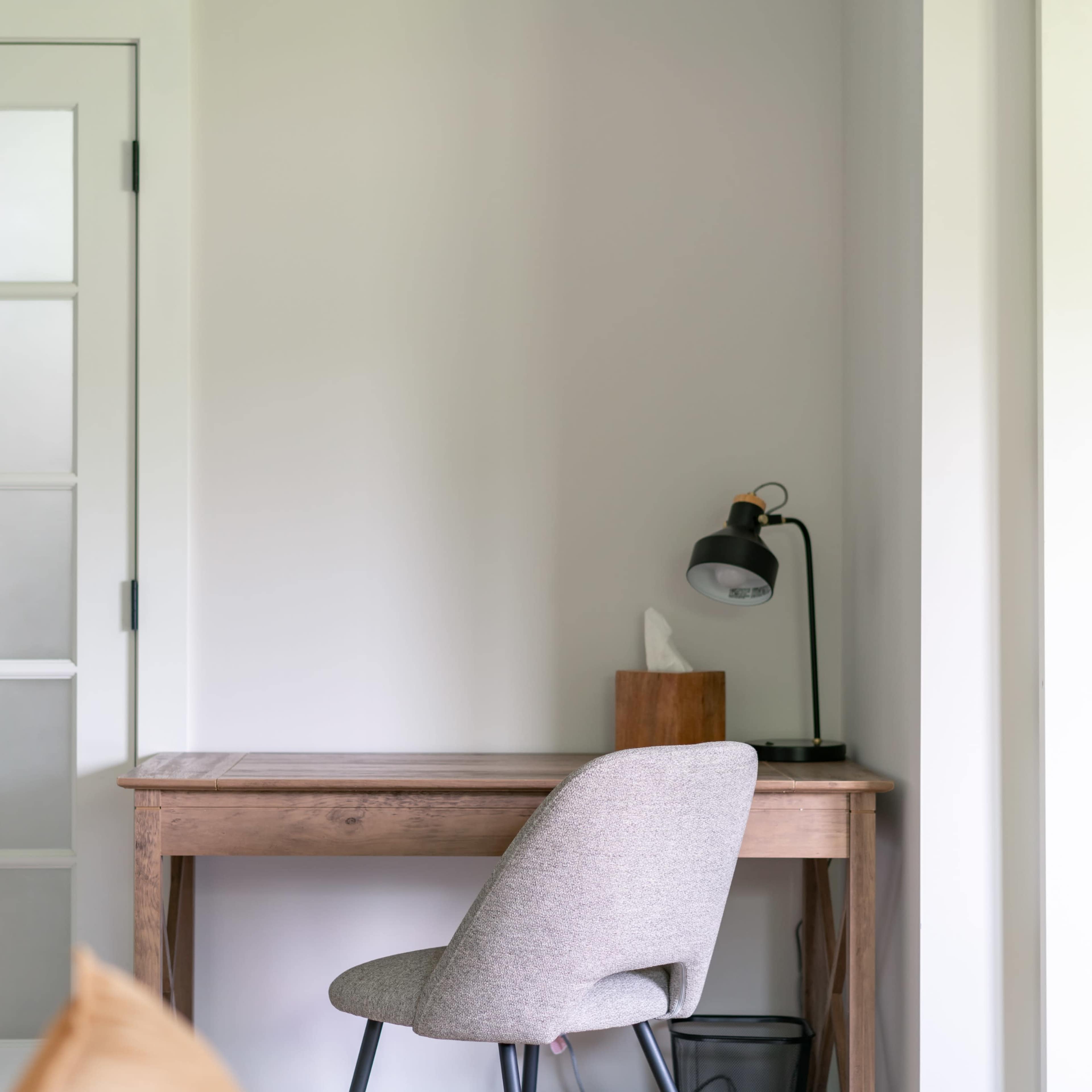 A wooden desk with a gray chair is positioned against a light wall beside a door, featuring a desk lamp and a wooden box.